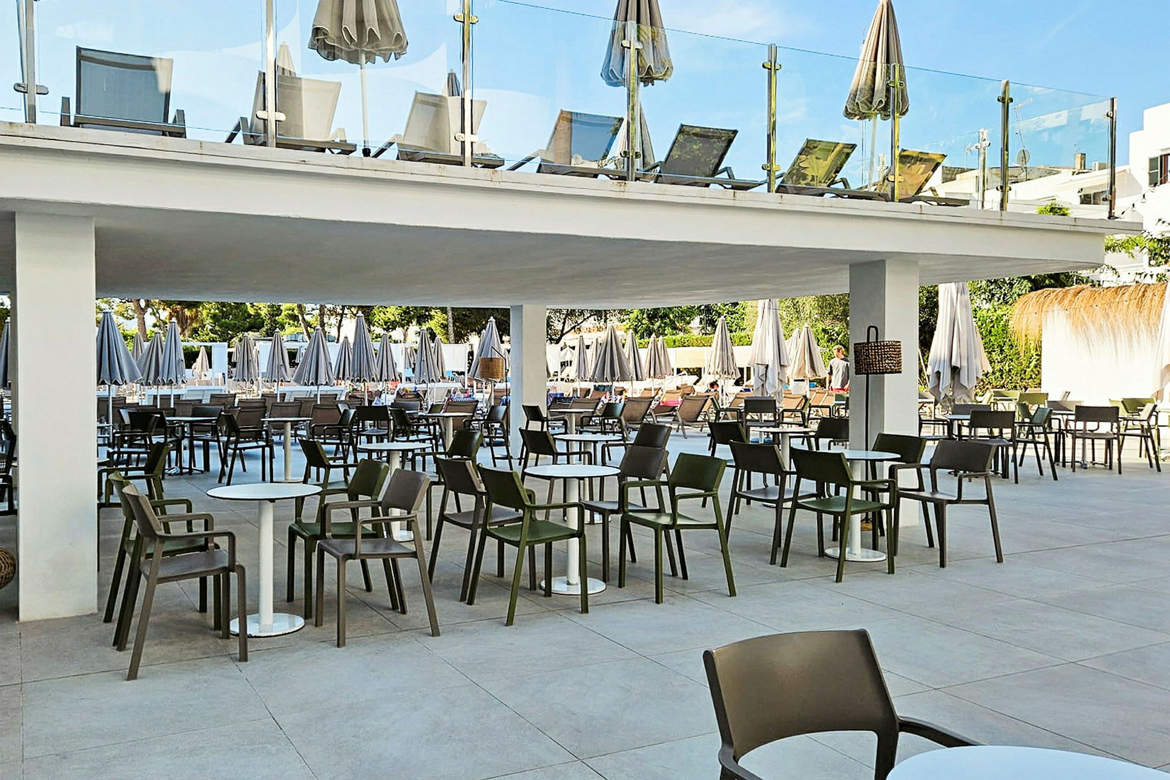 Billede af en terrasse med borde og stole under et tag samt liggestole på en etage ovenover. (AI-genereret tekst)