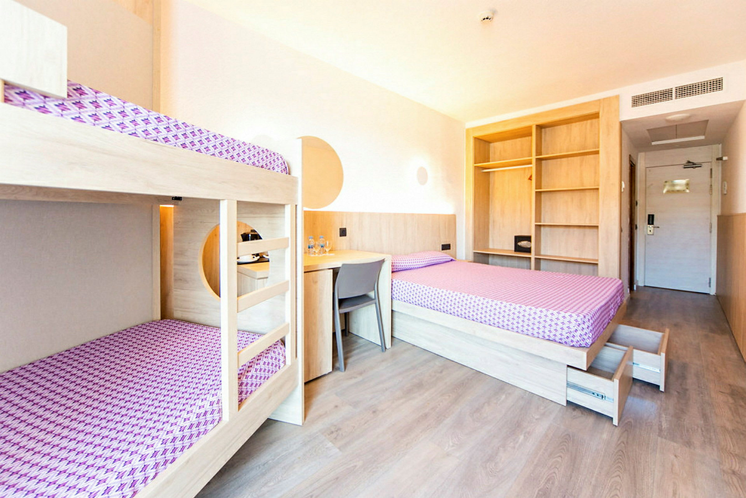 Et hotelværelse med køjesenge, en enkeltseng, skrivebord og åbne hylder. Et dørområde kan ses i baggrunden. (AI-genereret tekst)