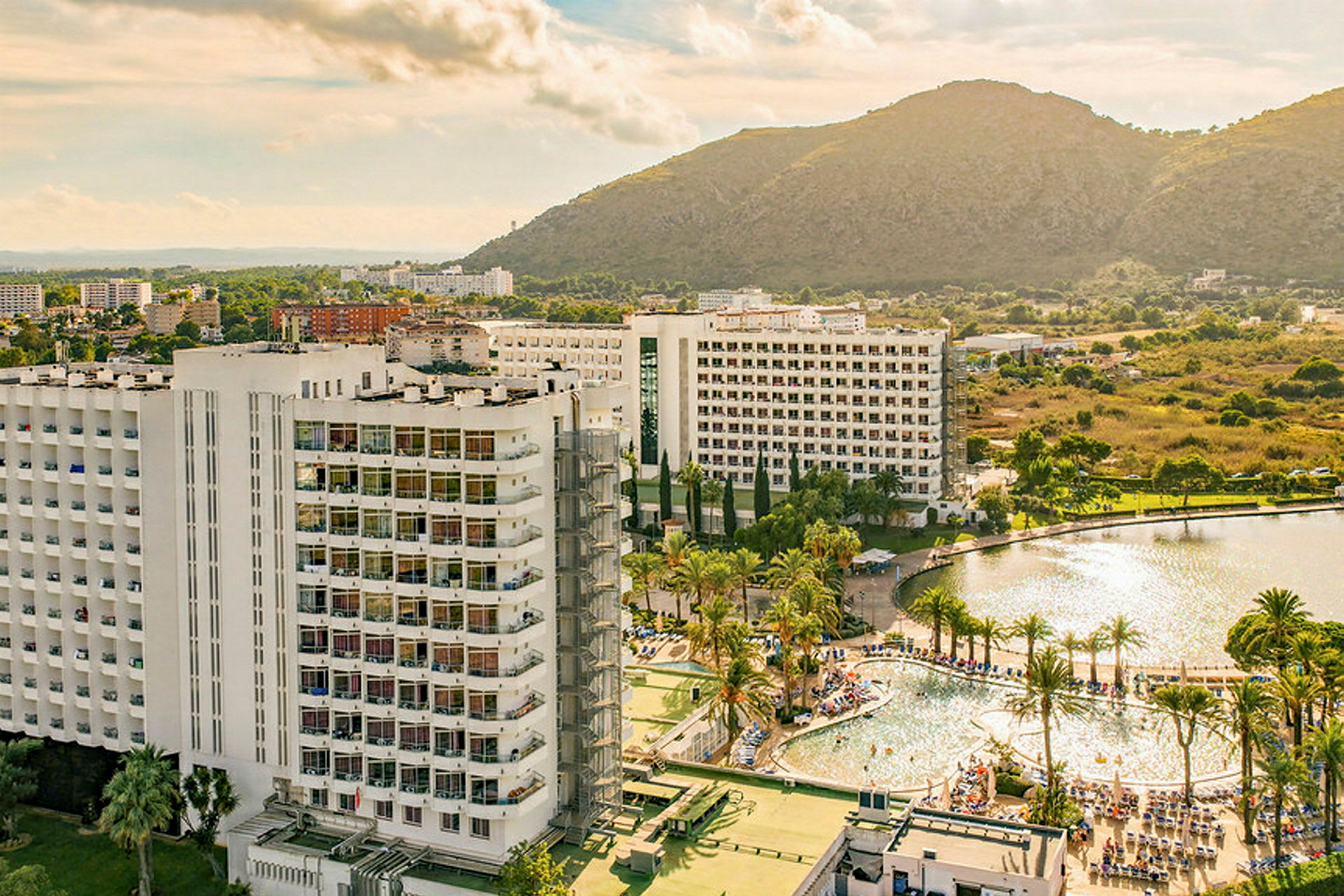 Billede af et hotel med omkringliggende poolområde, palmer og bjerge i baggrunden. (AI-genereret tekst)
