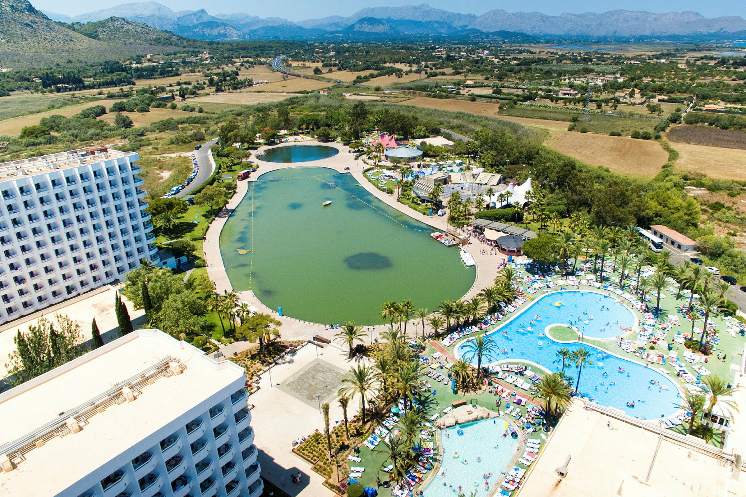 Luftfoto af et feriekompleks med hotelbygninger, pools og en kunstig sø omgivet af grønne områder og landskab. (AI-genereret tekst)