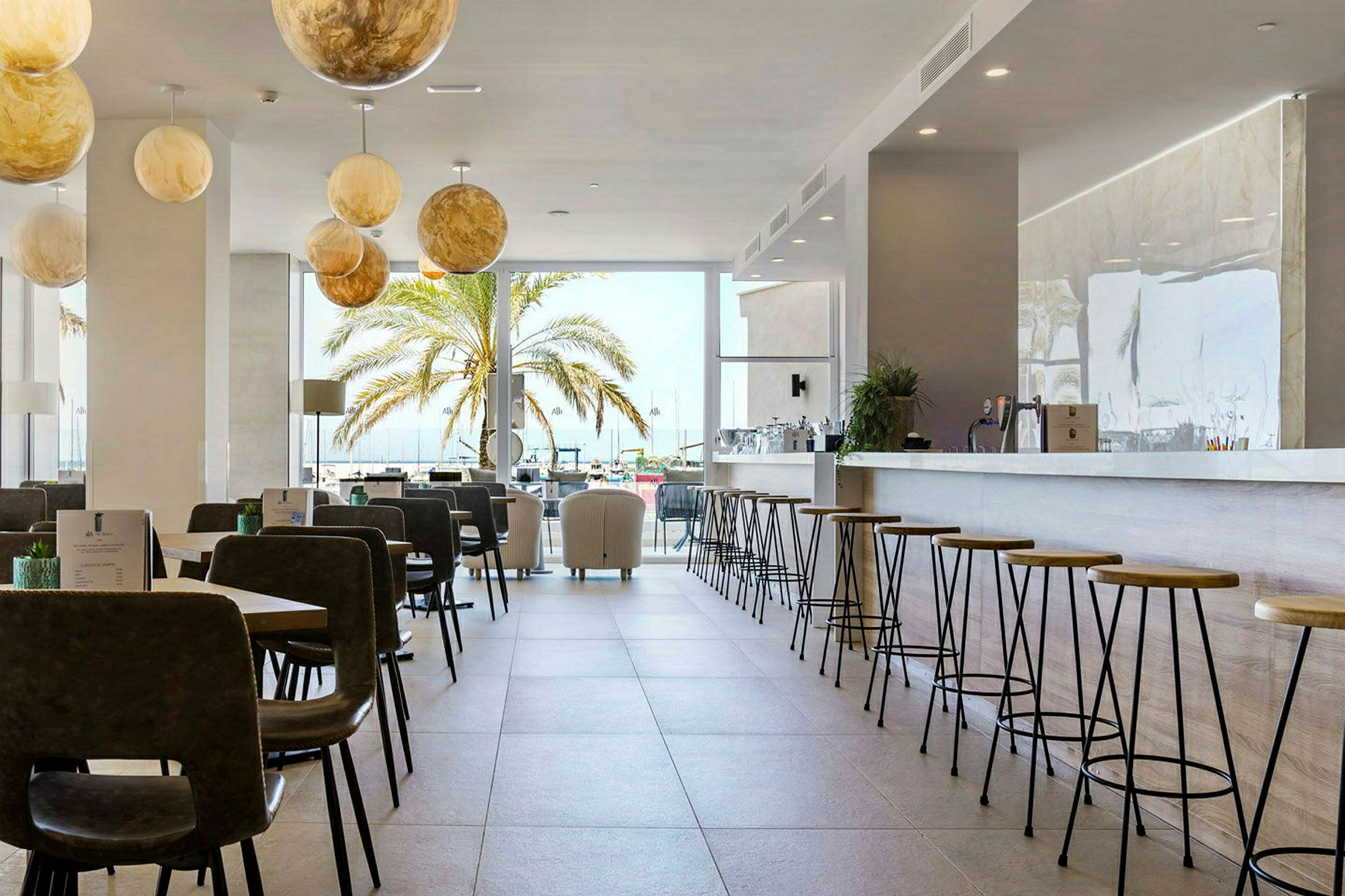 En lys bar og restaurant med siddepladser samt en palme udenfor et stort vindue. (AI-genereret tekst)