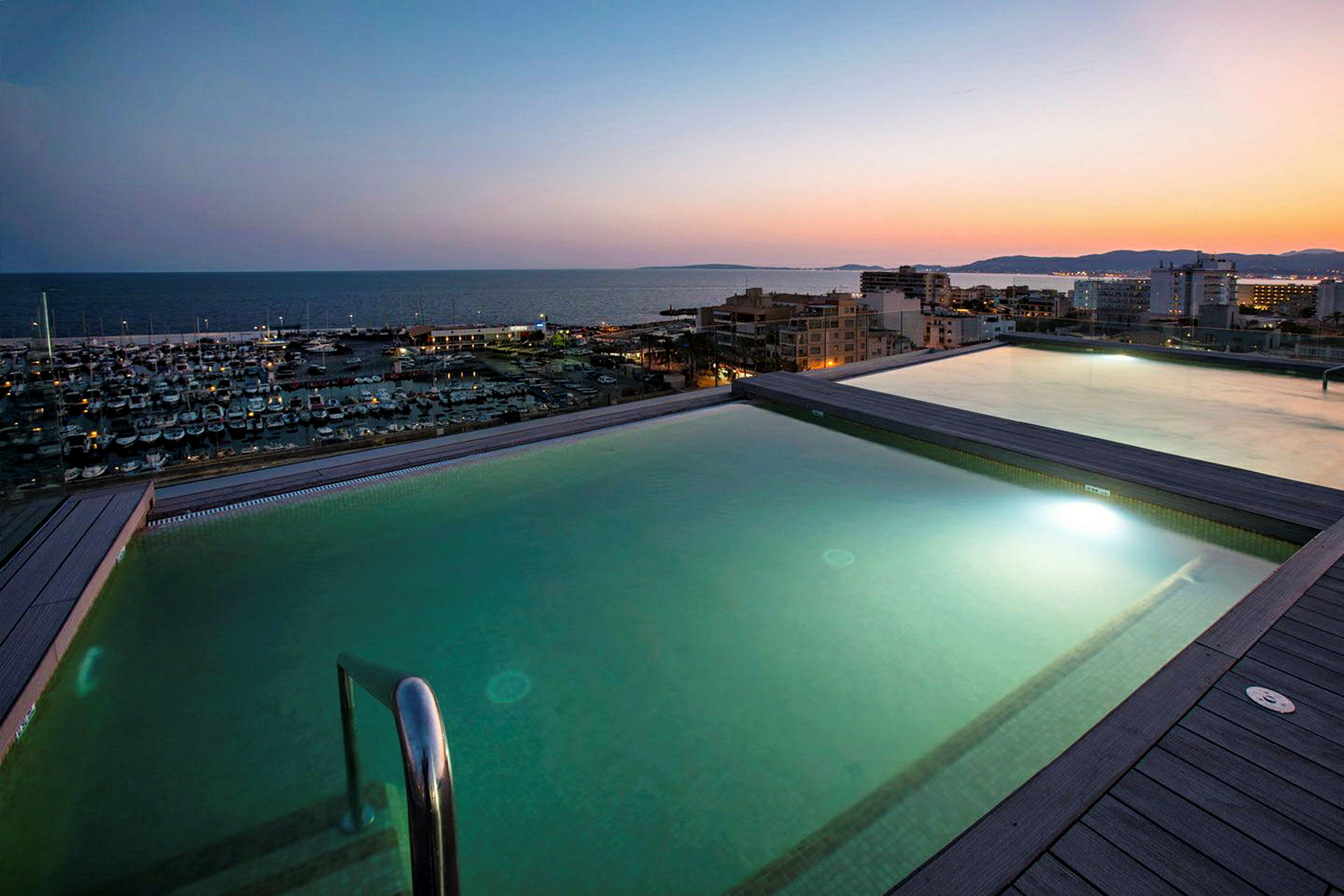 To oplyste pools med udsigt over en lystbådehavn og havet ved solnedgang. (AI-genereret tekst)