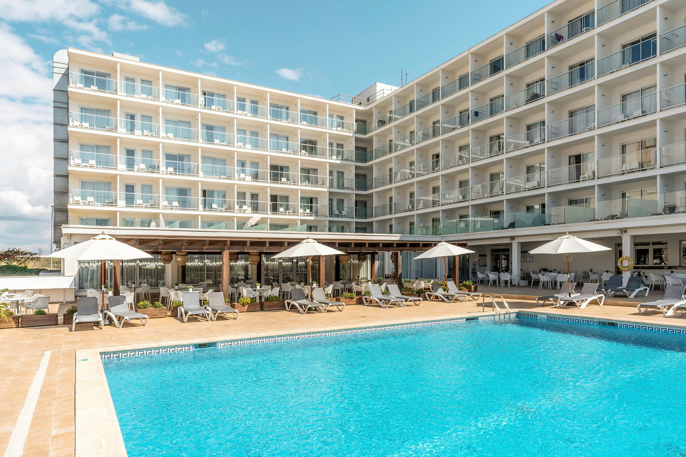 Et hotel med flere etager og altaner. Foran bygningen er der en udendørs pool, liggestole og parasoller. (AI-genereret tekst)