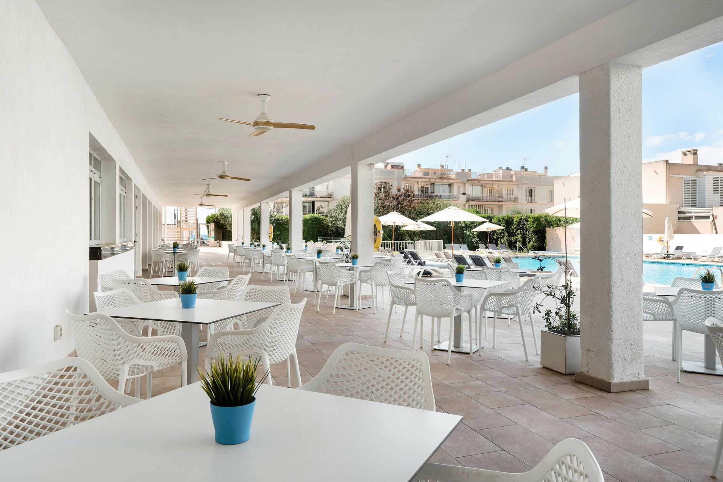 En udendørs terrasse med hvide borde og stole samt små planter i potter på bordene. I baggrunden ses en pool og liggestole med parasoller. (AI-genereret tekst)
