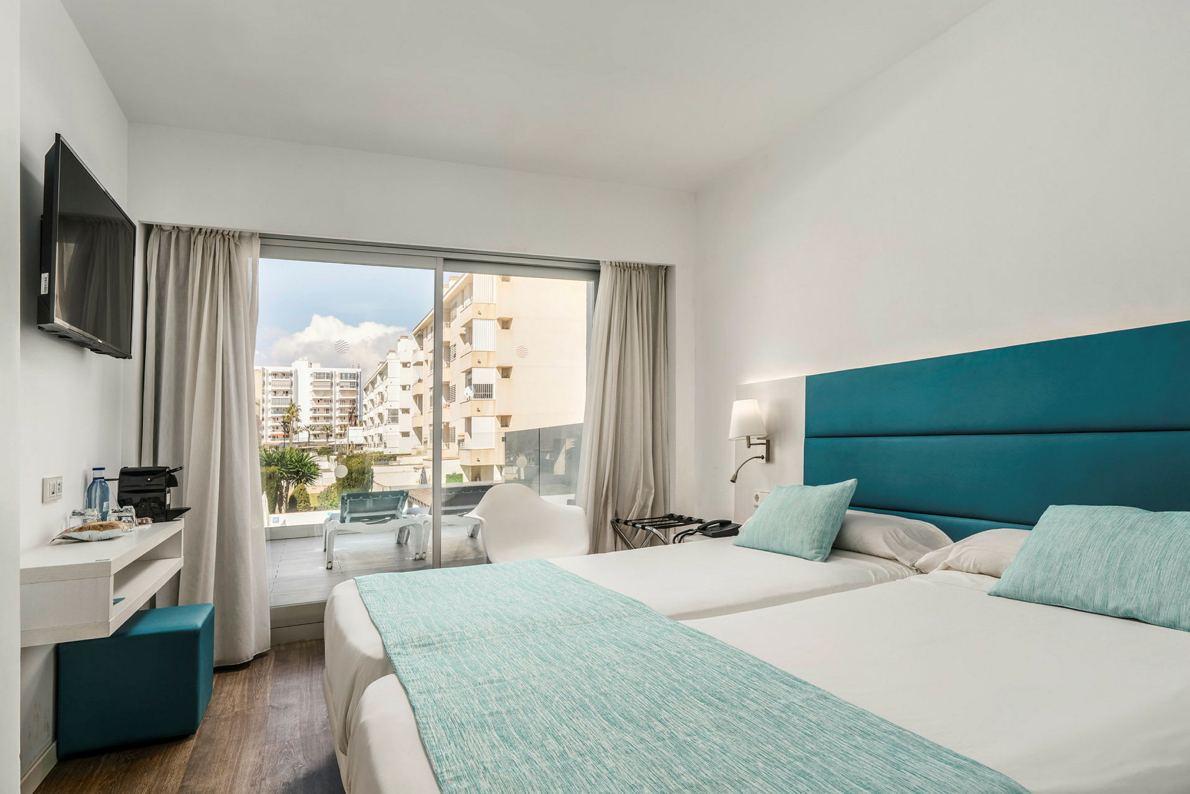 Billede af et hotelværelse med to enkeltsenge ved siden af hinanden, turkis sengetøj og puder samt moderne indretning. En glasdør giver udsigt til en altan og et område med bygninger. (AI-genereret tekst)