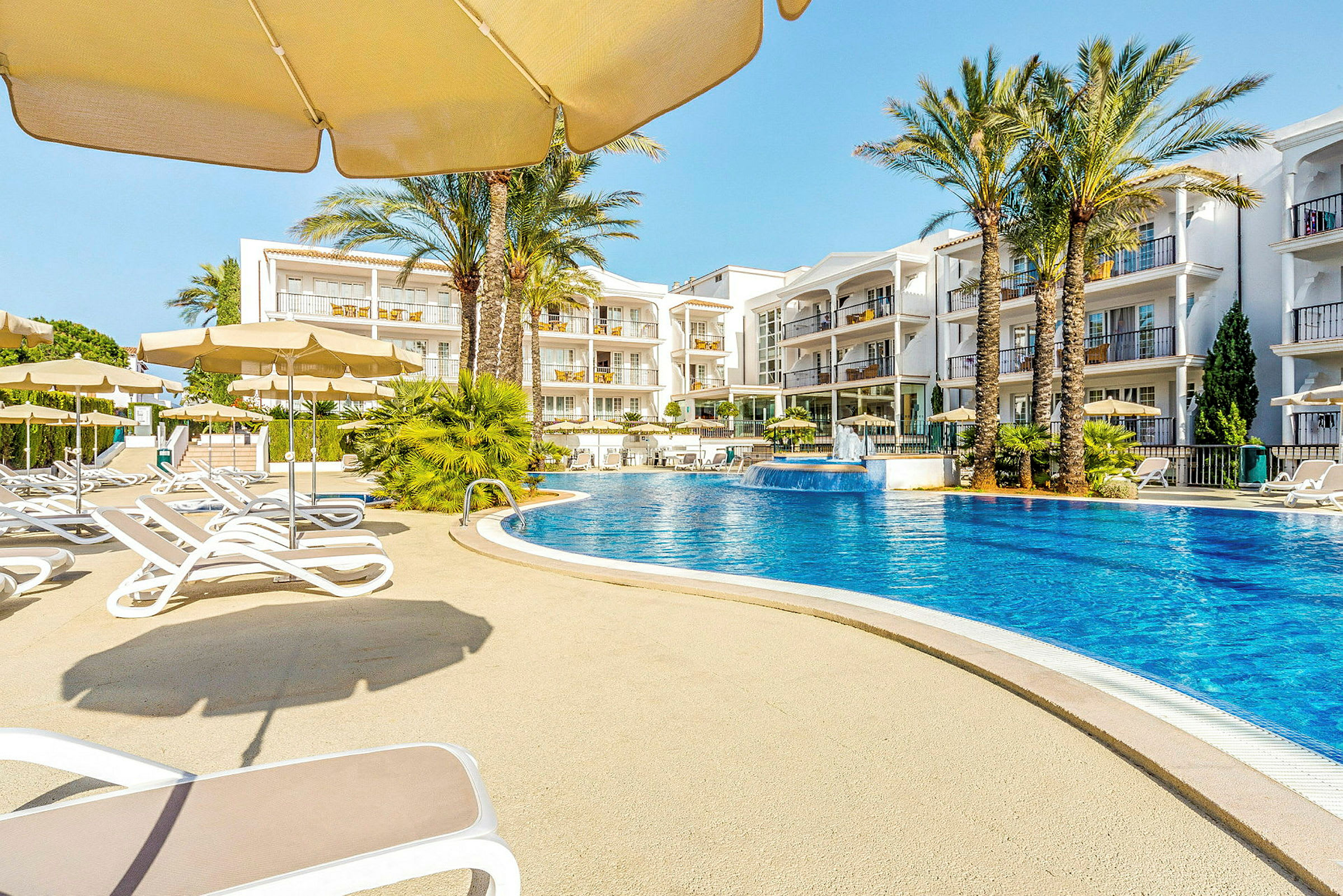 En udendørs pool omgivet af liggestole og parasoller med hotelbygninger i baggrunden. (AI-genereret tekst)