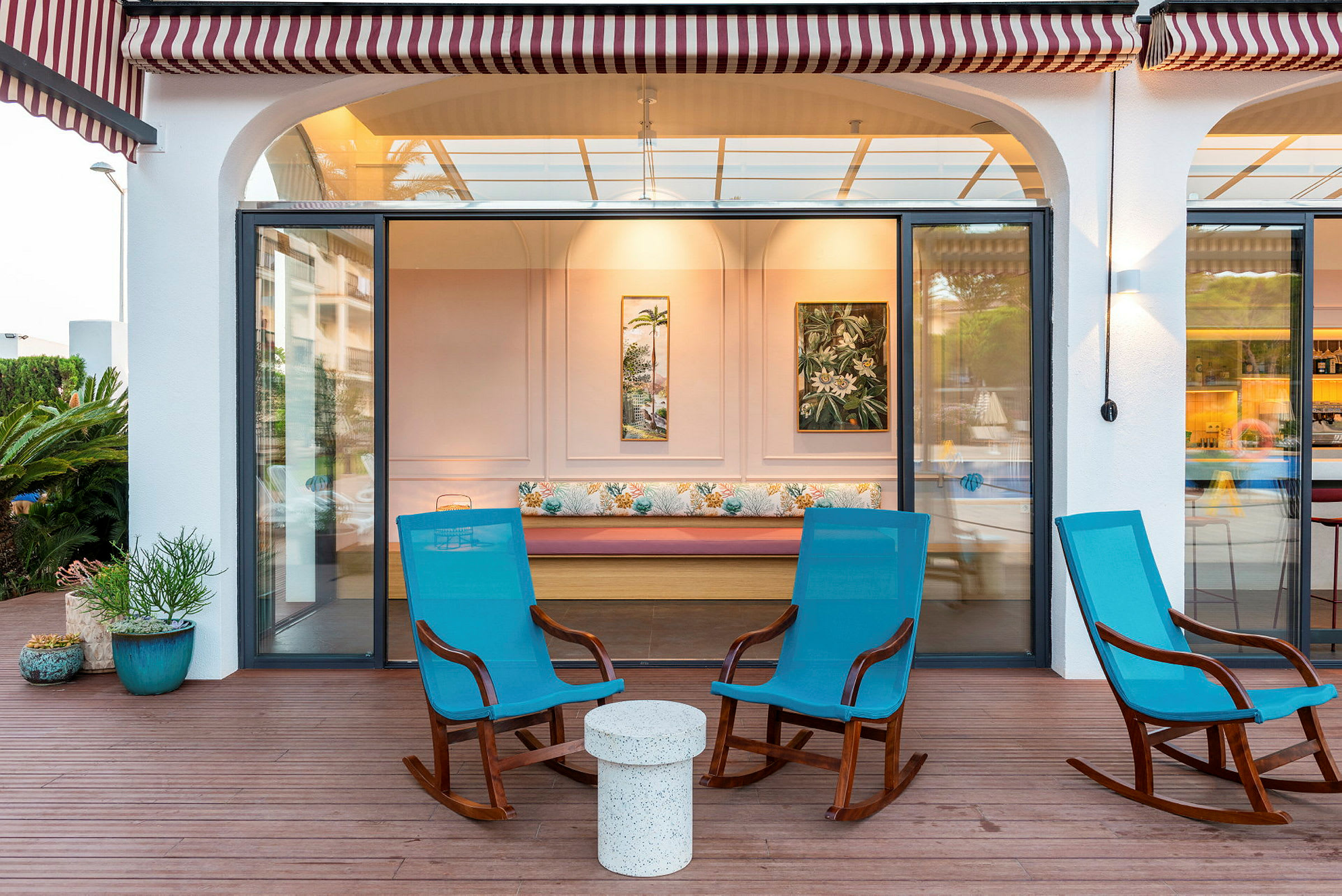 Billede af en terrasse med tre blå gyngestole og et lille bord foran en bygning med glasdøre. (AI-genereret tekst)