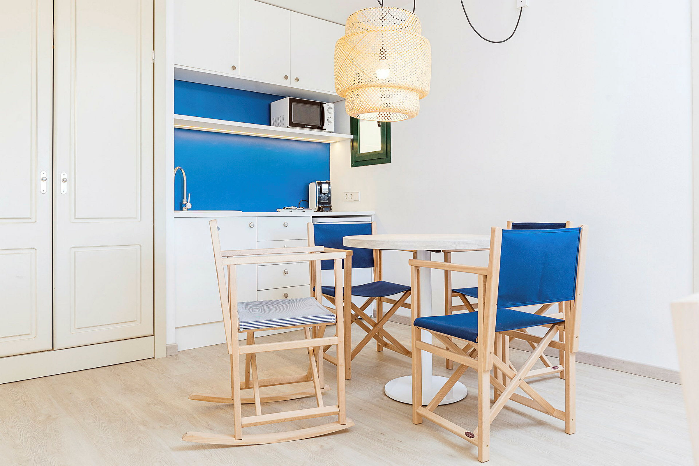 Et lille køkken med blå og hvide overflader, en spisegruppe med træstole og et rundt hvidt bord. En loftslampe med flettet design hænger over bordet. (AI-genereret tekst)