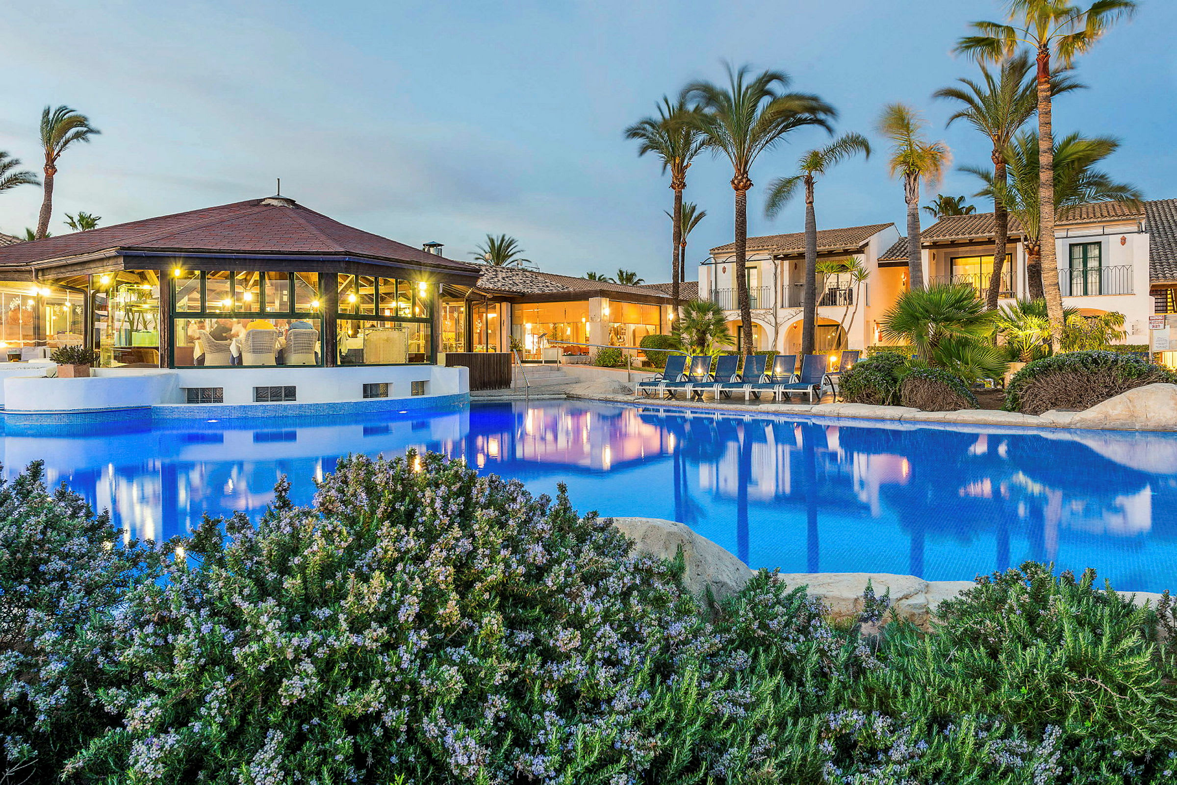 En hotelbygning med pool og liggestole omgivet af palmer. (AI-genereret tekst)