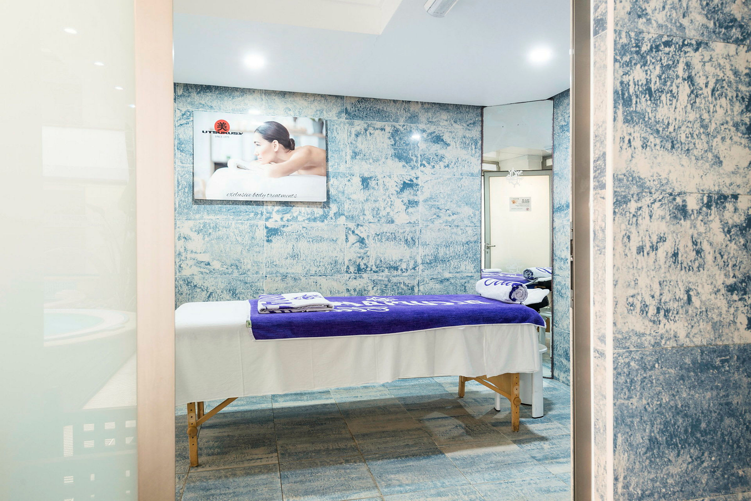 Et spa-rum med massagebænk dækket af hvide og lilla håndklæder, blå mosaik vægdesign og spejl. (AI-genereret tekst)
