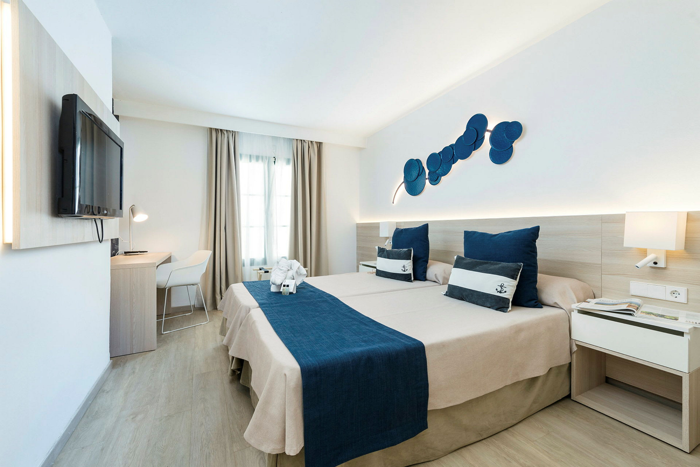 Hotelværelse med to senge og maritimt tema, blåt sengetøj og dekorative puder. (AI-genereret tekst)