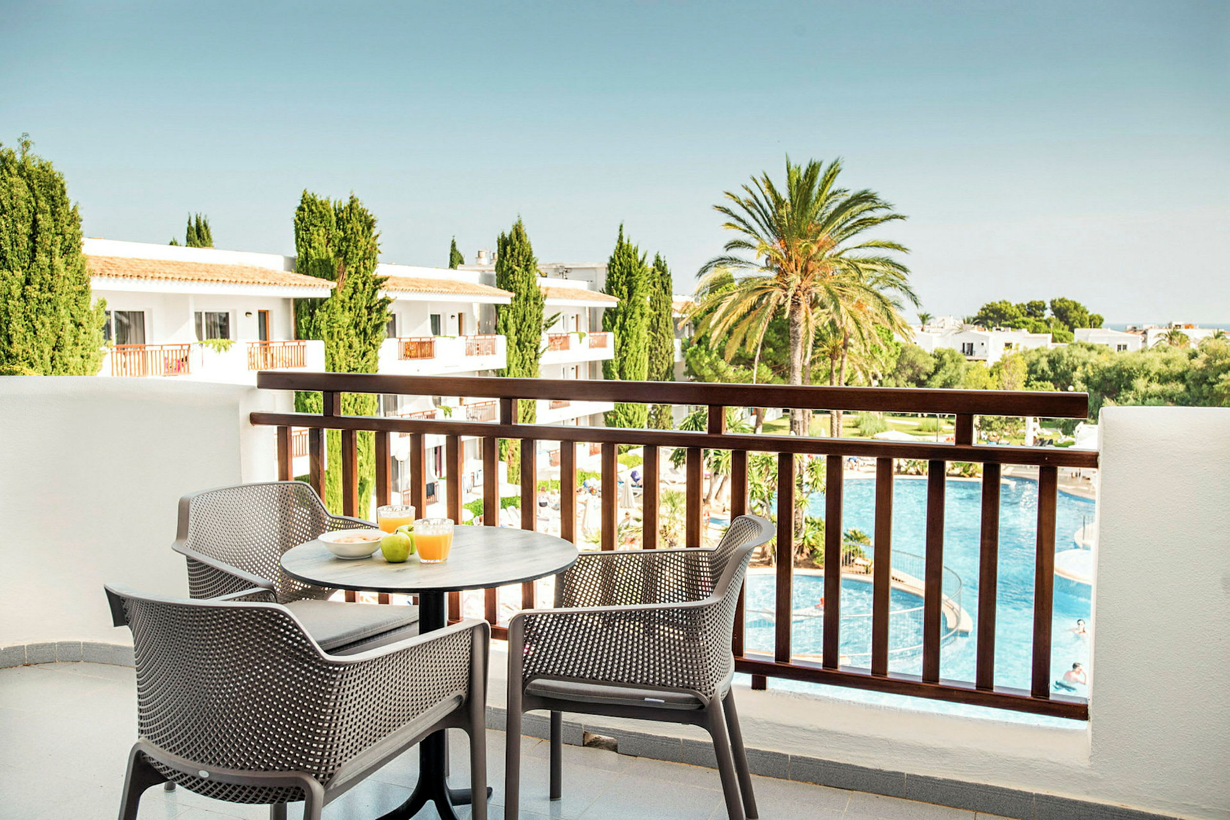 Udsigt fra en balkon med et bord og stole, hvor morgenmad er fremstillet. I baggrunden er der en pool, grøntområder og bygninger. (AI-genereret tekst)