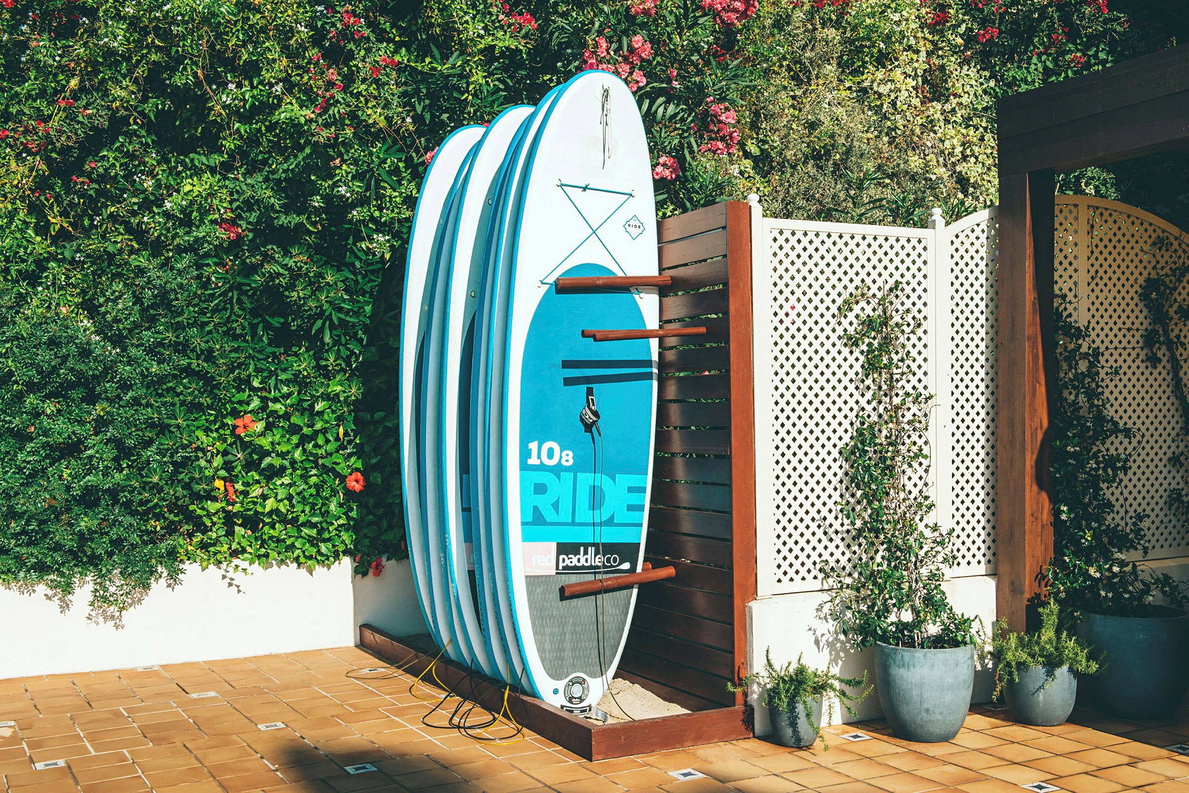 Billedet viser et sæt blå paddleboards, der læner op ad et træstativ på en solrig terrasse. I baggrunden er frodige grønne planter og blomster. (AI-genereret tekst)