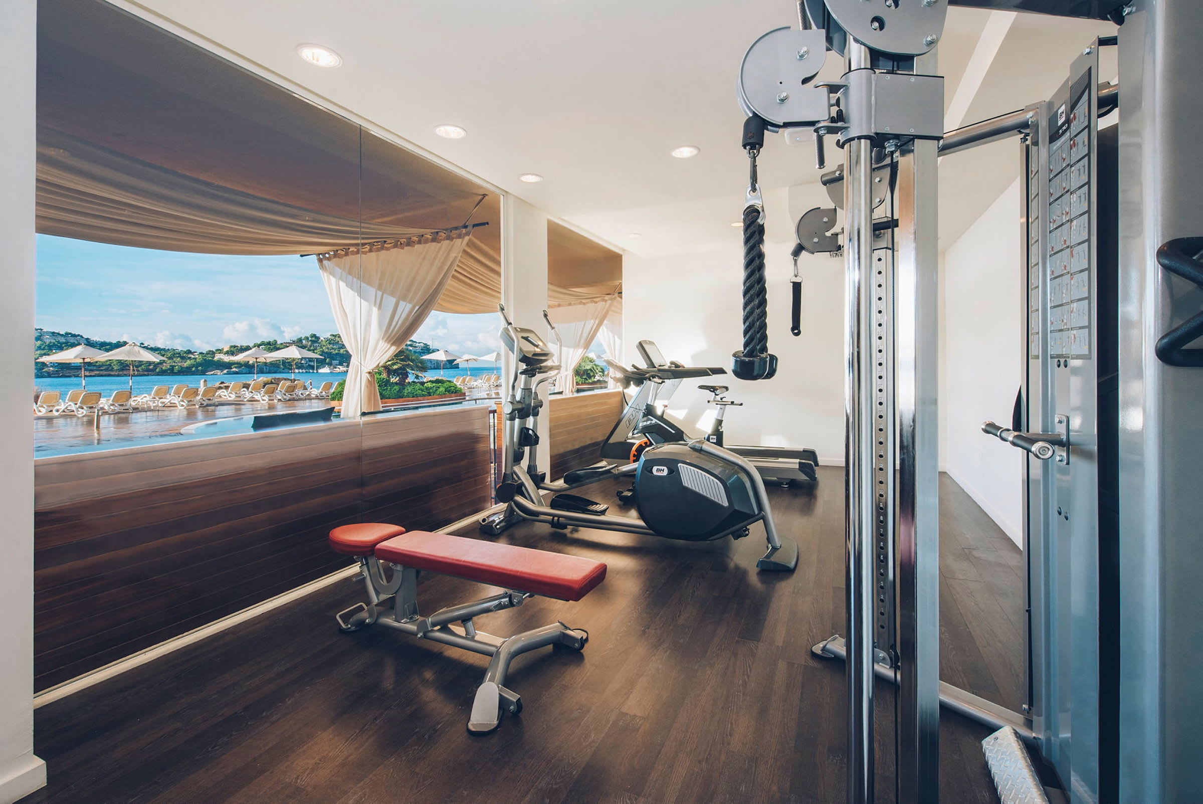 Et fitnessrum med træningsmaskiner og en rød træningsbænk, med udsigt til en pool og liggestole gennem store glasvinduer. (AI-genereret tekst)