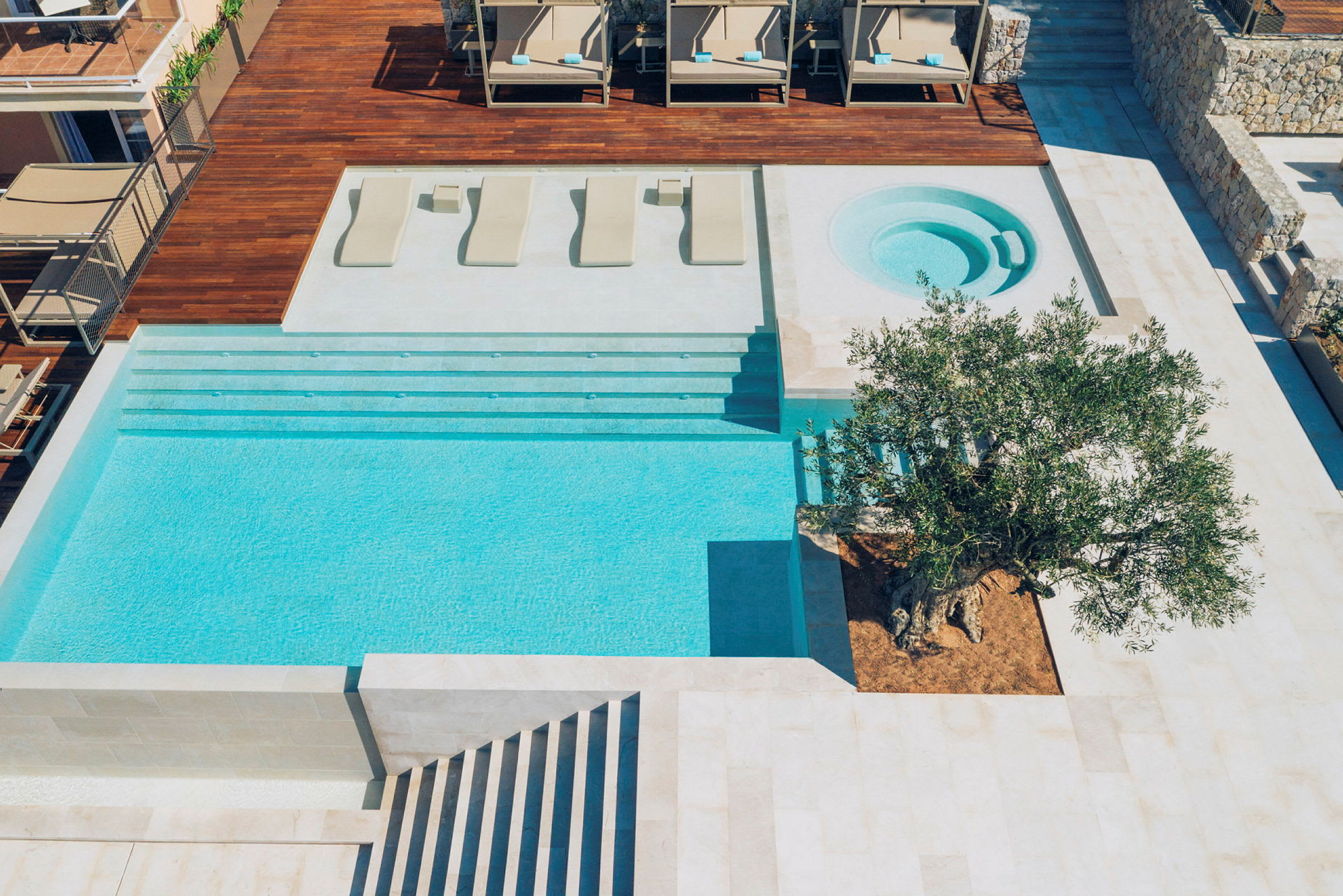 En moderne pool med tilhørende spabad og træterrasse. På terrassen er der nogle liggestole, og der er et stort oliventræ ved poolens kant. (AI-genereret tekst)
