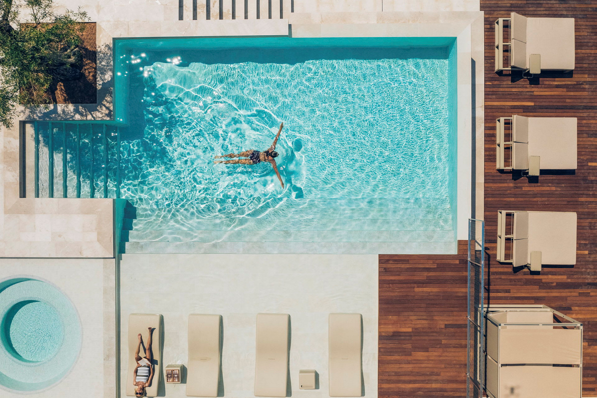 Billedet viser et overblik over en udendørs terrasse med en pool. I poolen svømmer en person, mens andre liggestole er placeret ved siden af på trædækket. (AI-genereret tekst)