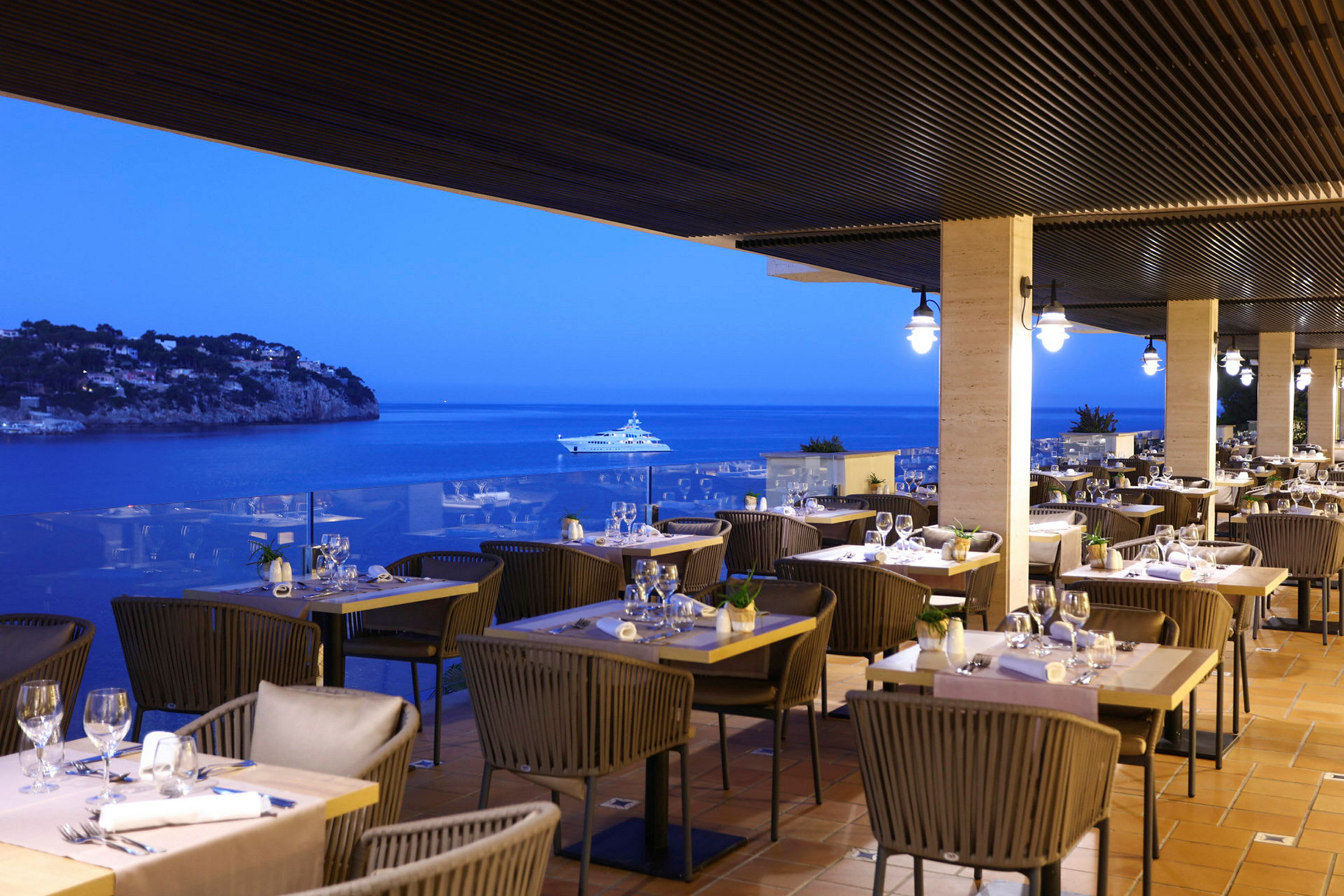 En restaurant med udendørs servering på en terrasse med udsigt over havet om aftenen. Flere borde er dækket op med stole og glas. (AI-genereret tekst)