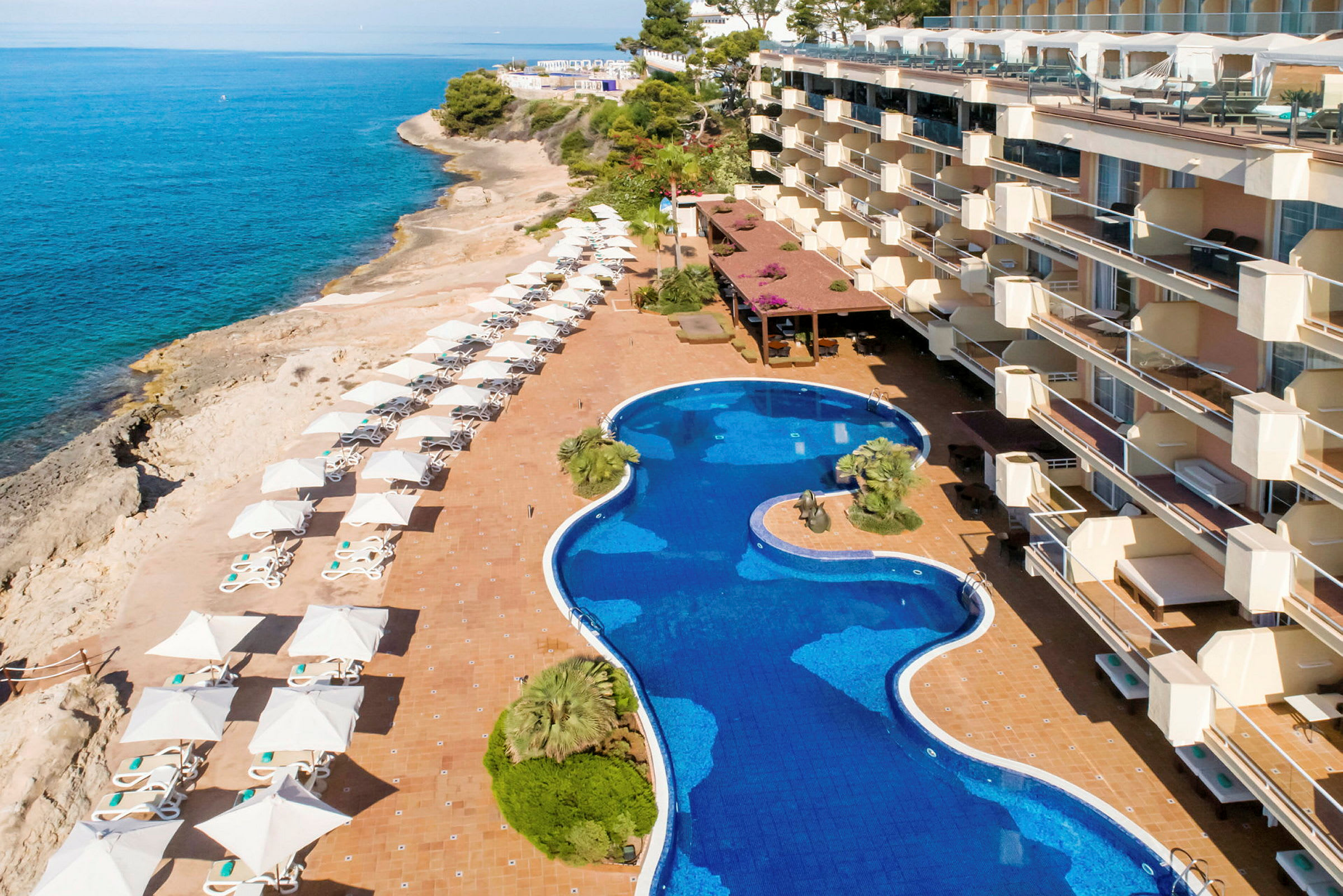 Billede af en hotelpool omgivet af liggestole og parasoller med udsigt over klipper og hav. (AI-genereret tekst)