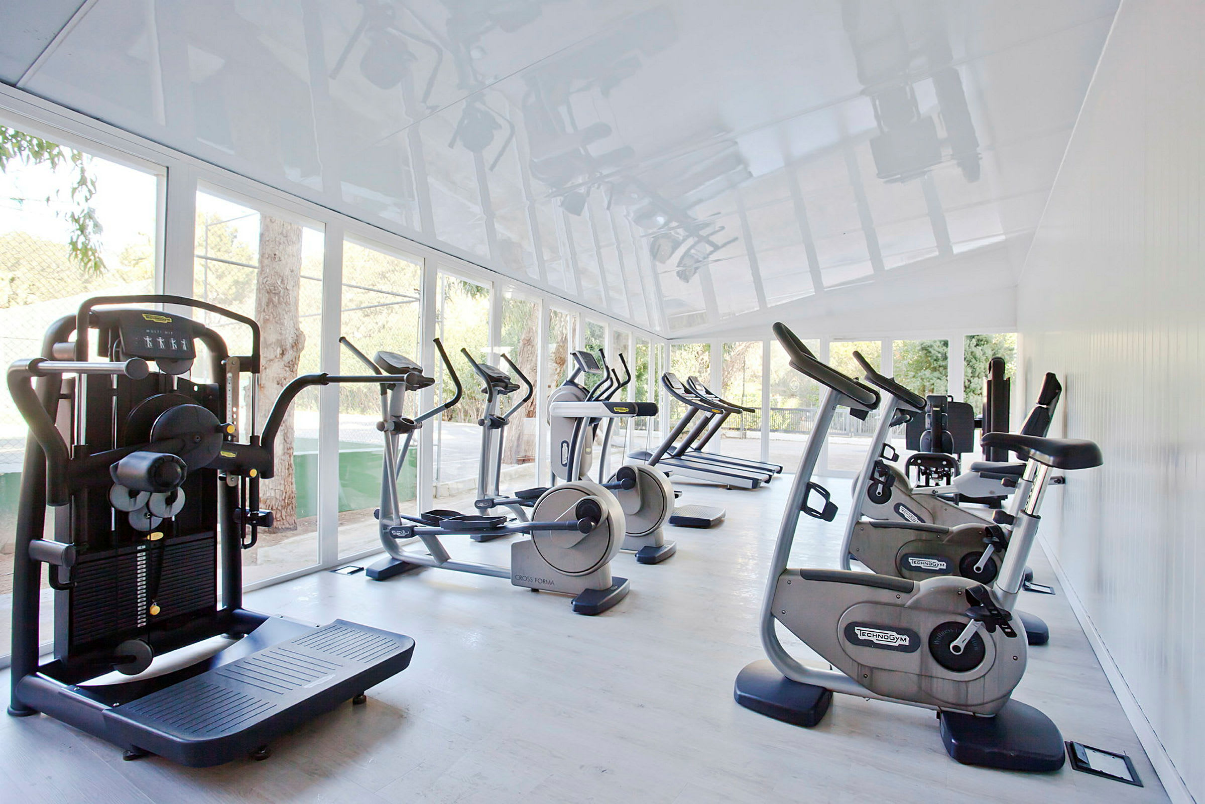 Et fitnesscenter med forskellige træningsmaskiner såsom kondicykler og løbebånd placeret i et lyst rum med store vinduer, der lukker naturligt lys ind. (AI-genereret tekst)