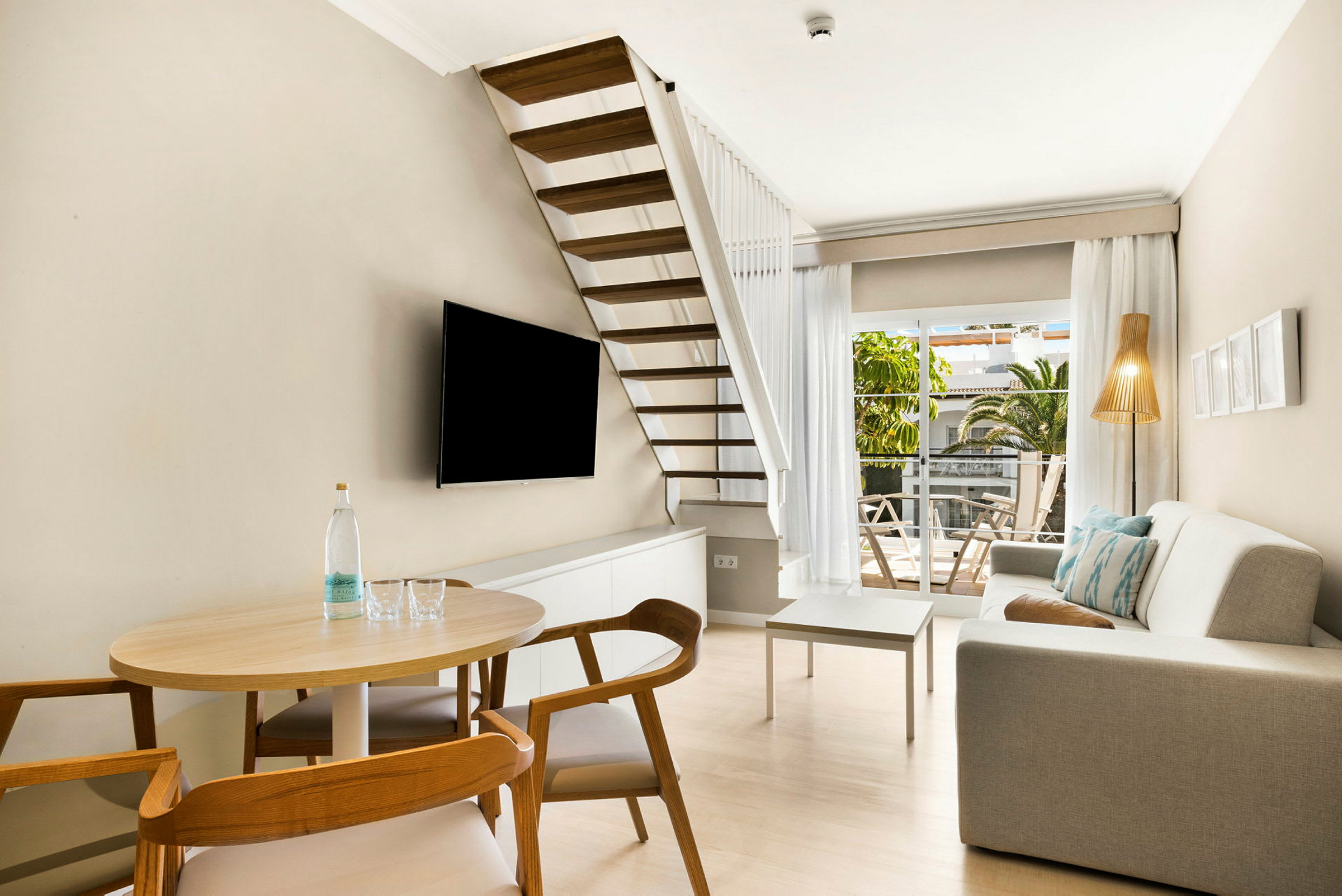 Moderne og lyst stue med trappe, sofa, TV og udsigt til en solrig terrasse. (AI-genereret tekst)