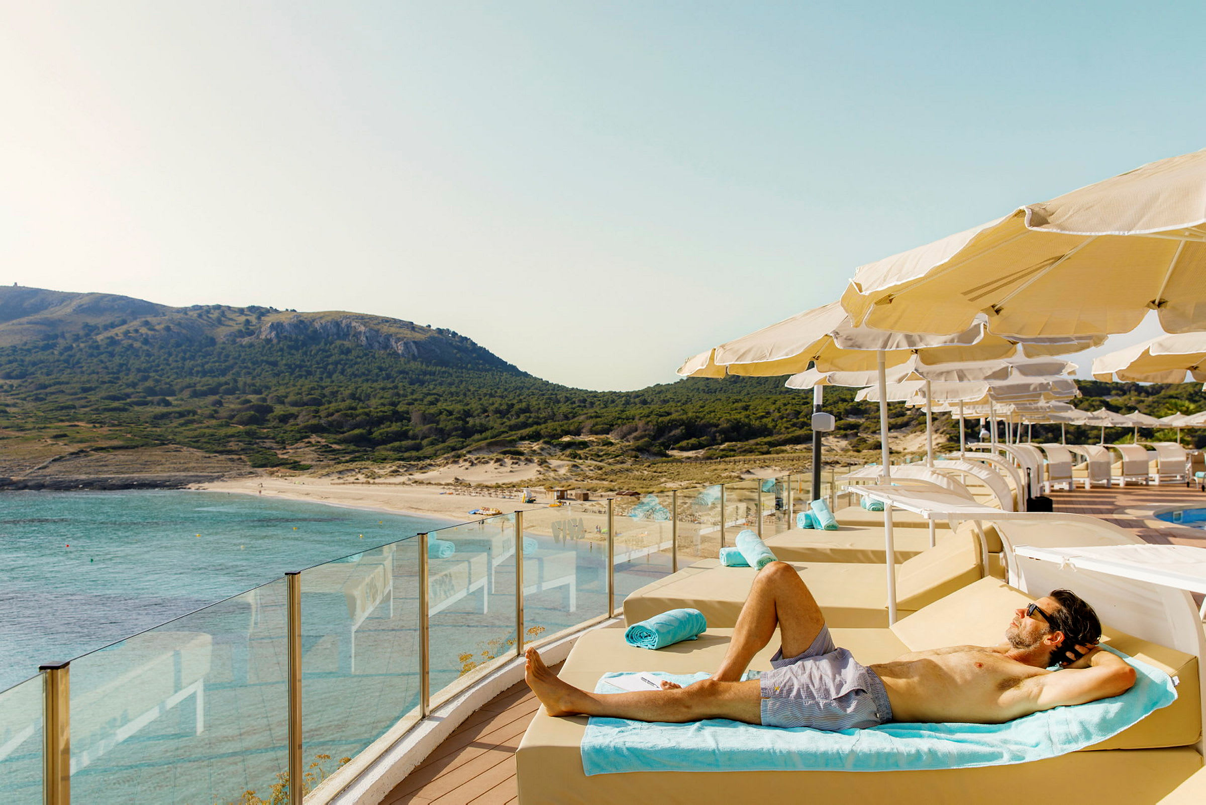 En person ligger på en solseng ved en pool med udsigt over en strand og bjergrigt landskab. (AI-genereret tekst)