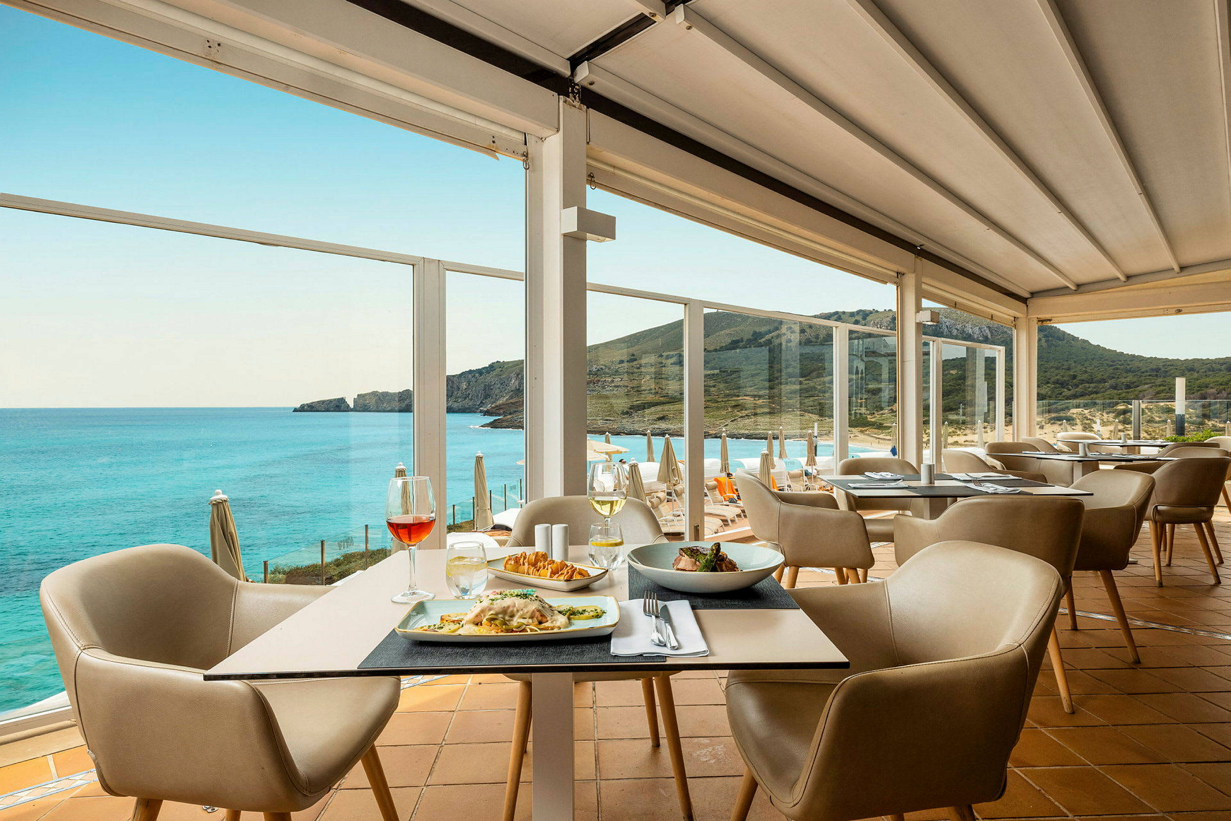 En restaurant med borde med mad og stole samt en panoramaudsigt over havet og en bjergkyst. (AI-genereret tekst)