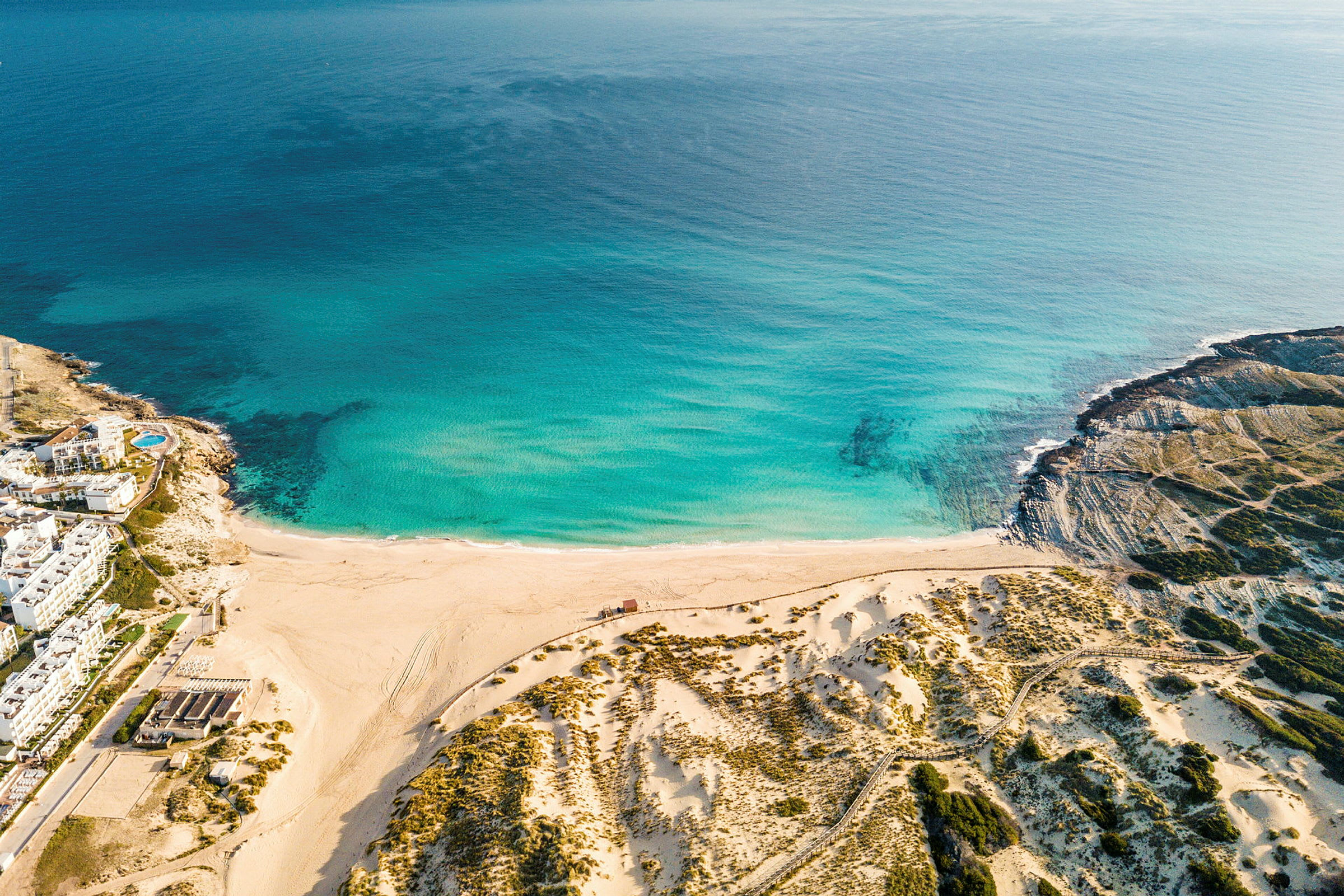 Luftfoto af en strand med turkis vand og sandklitter, omgivet af kystgrønne områder. (AI-genereret tekst)
