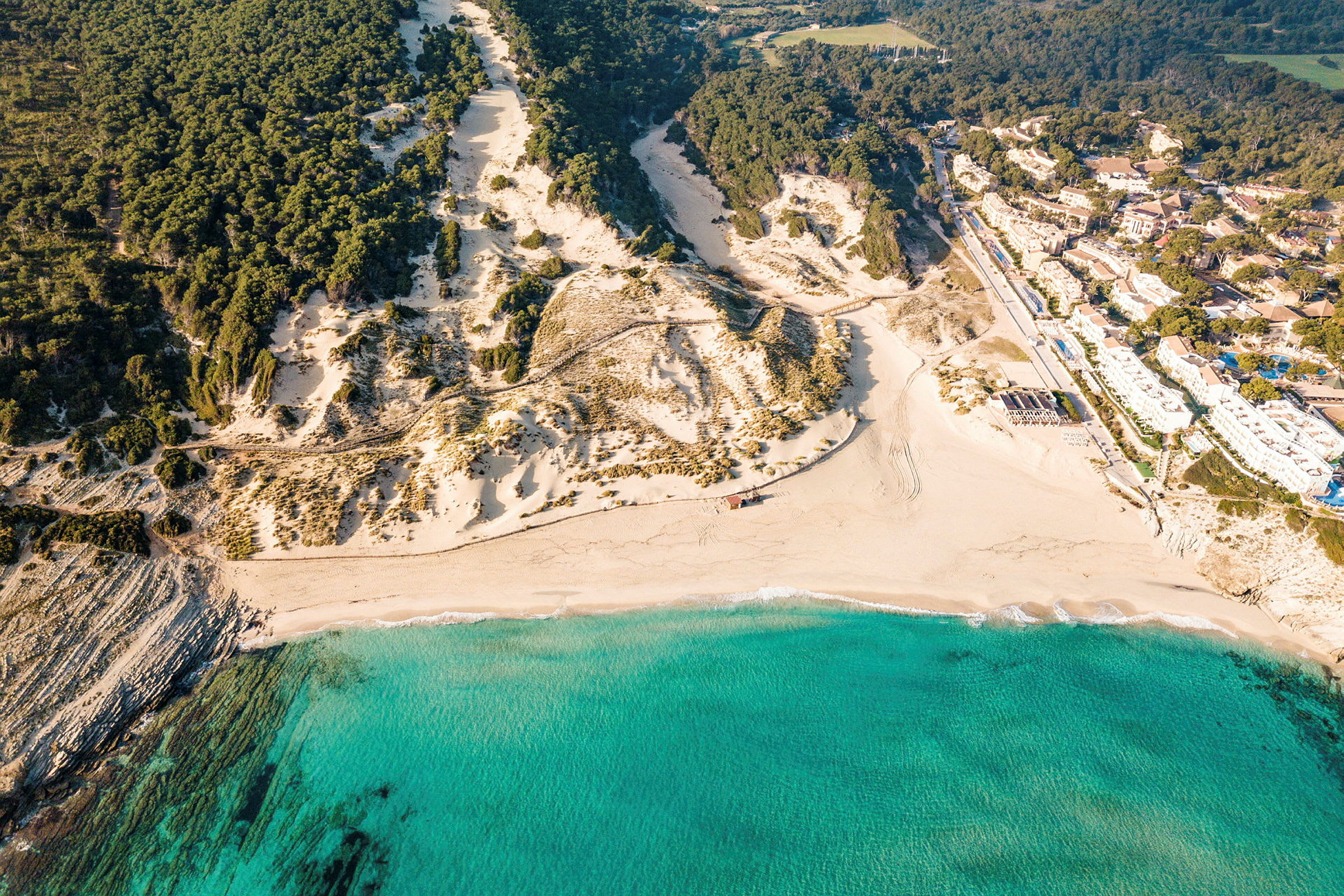 Luftfoto af en sandstrand med turkis vand, omgivet af klipper og fyrreskov. (AI-genereret tekst)
