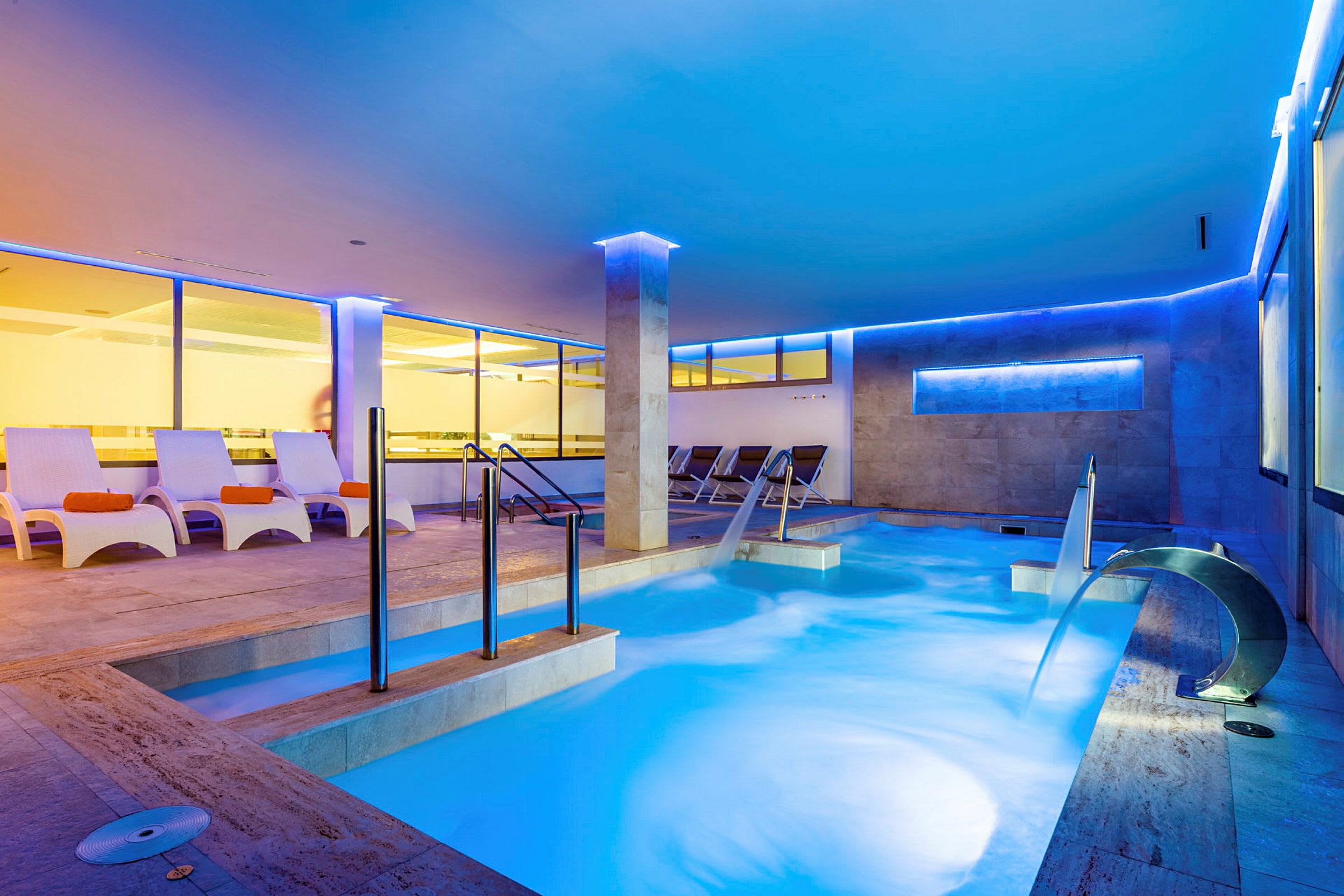 Indendørs pool med siddepladser og vandkaskade i et moderne oplyst spa. (AI-genereret tekst)