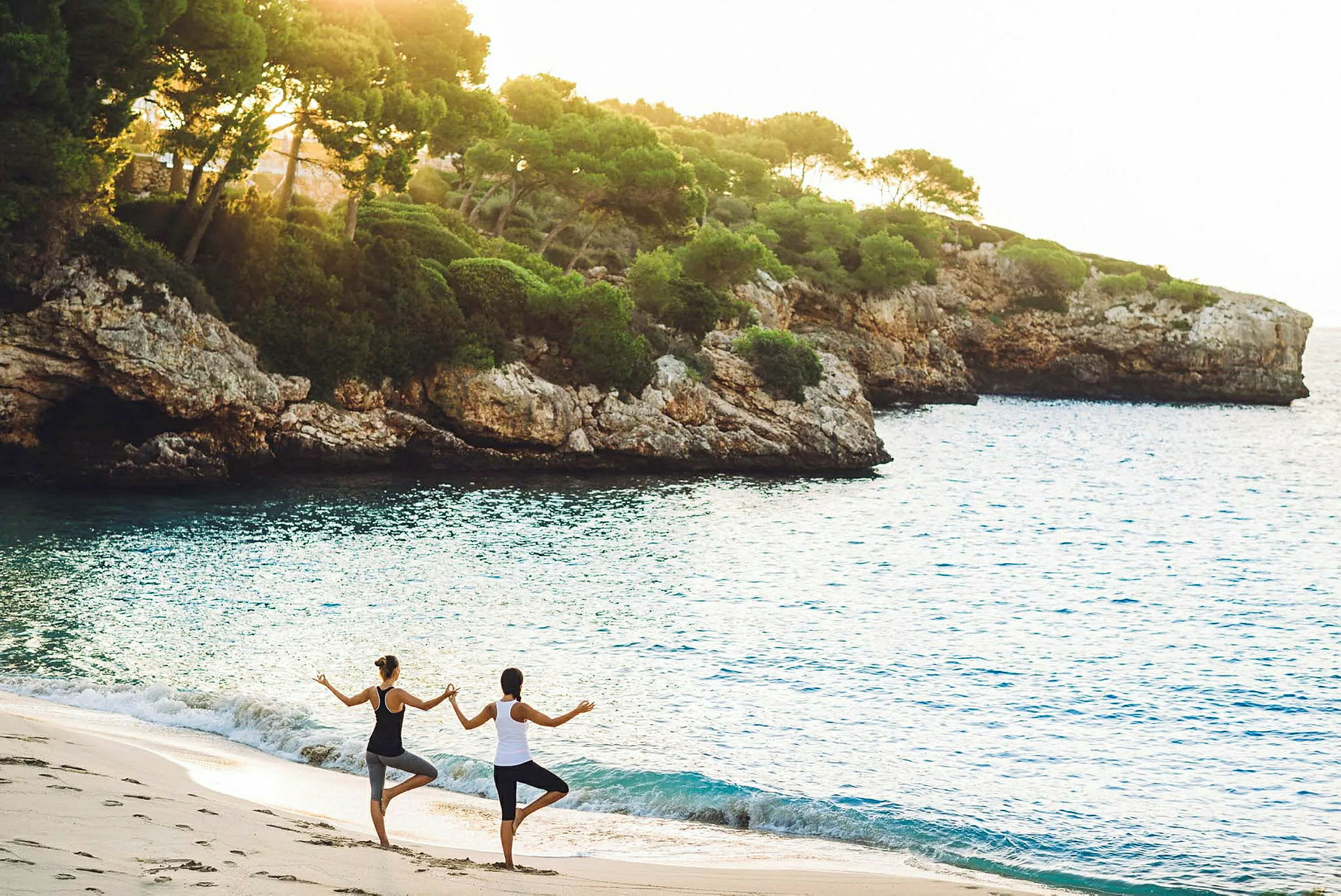 To personer står på en strand og udfører yoga i solnedgangen foran klipper og hav. (AI-genereret tekst)