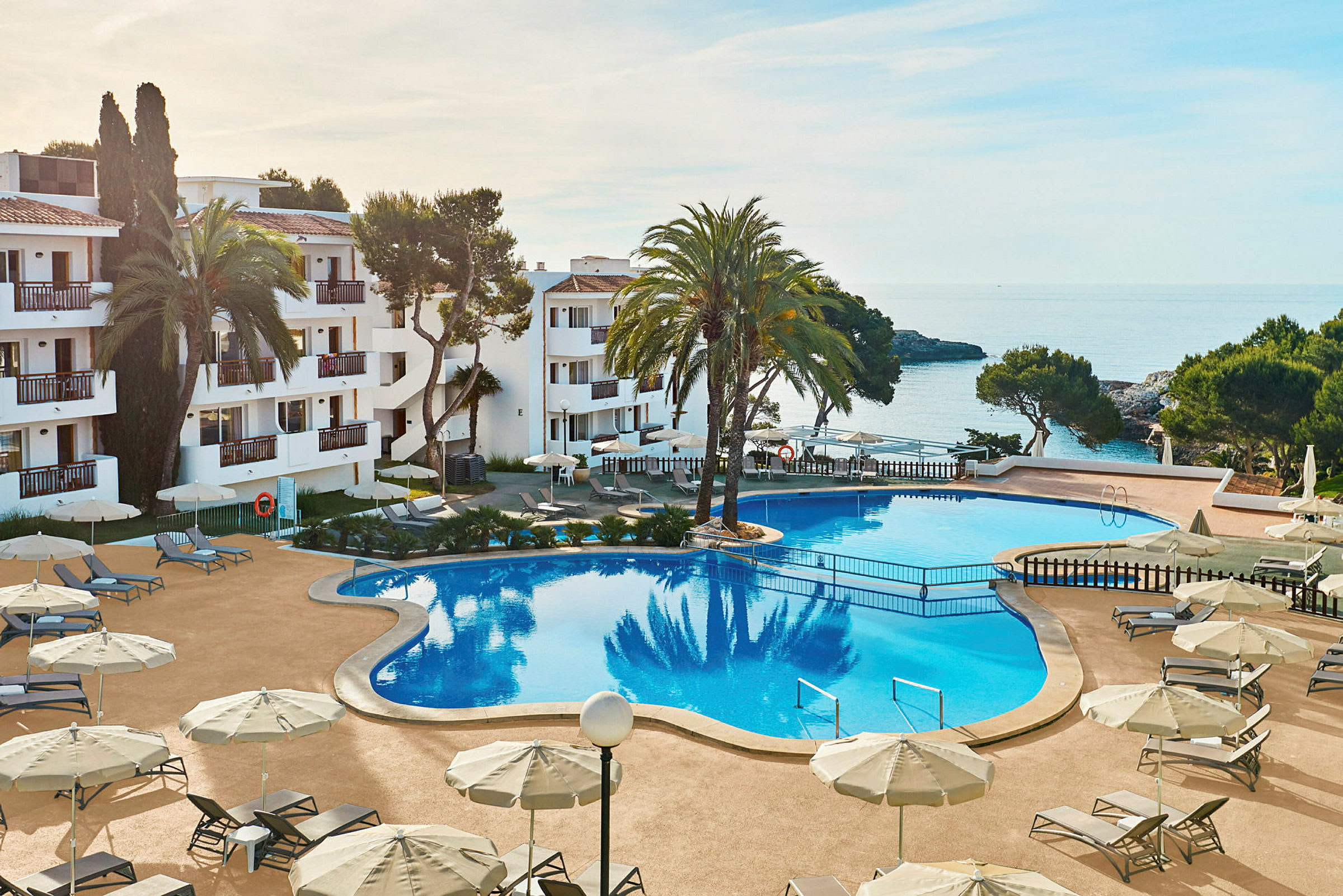 Billede af et hotelområde med en stor pool omgivet af liggestole og parasoller. I baggrunden ses havet og dele af bygningen. (AI-genereret tekst)