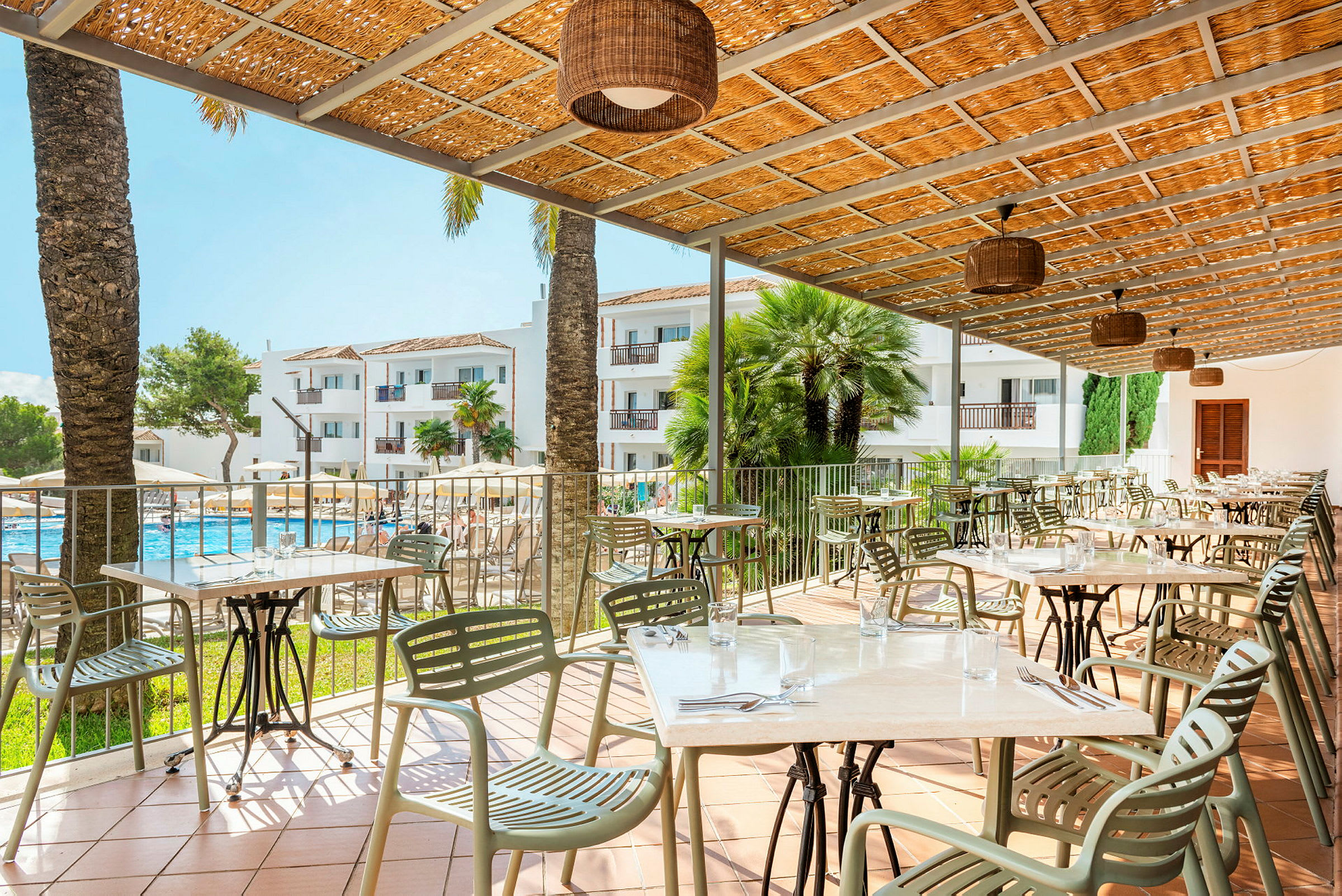 En udendørsrestaurant med borde og stole under et tag, med udsigt over en pool og hotelbygninger i baggrunden. (AI-genereret tekst)