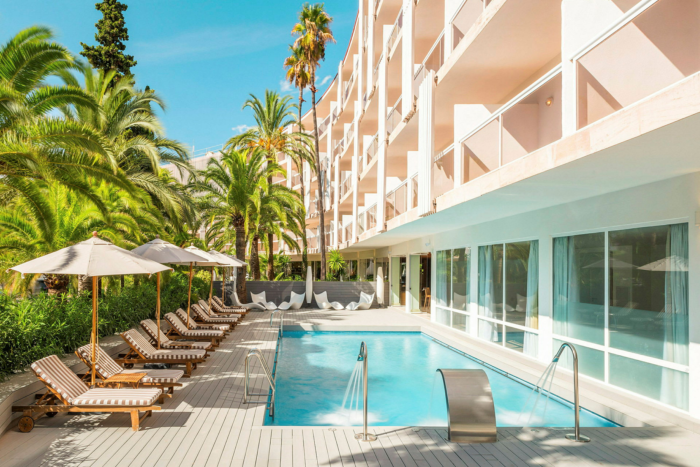 Udendørs pool omgivet af liggestole og parasoller, ved siden af hotelbygning med altaner. (AI-genereret tekst)