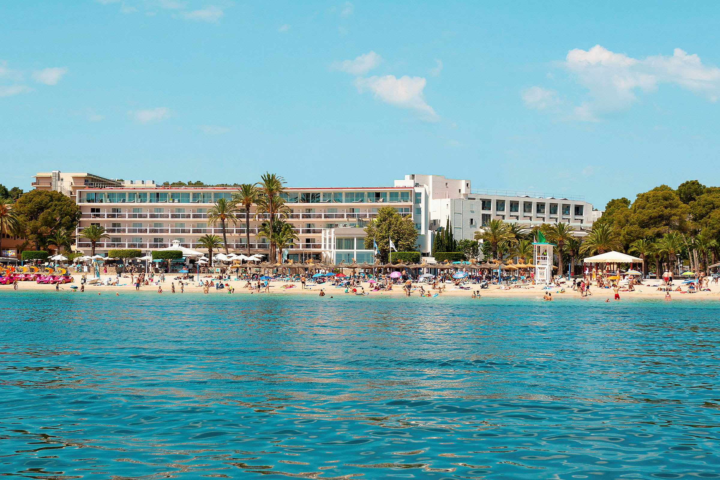 Et hotel ved en strand med palmer og badegæster. (AI-genereret tekst)