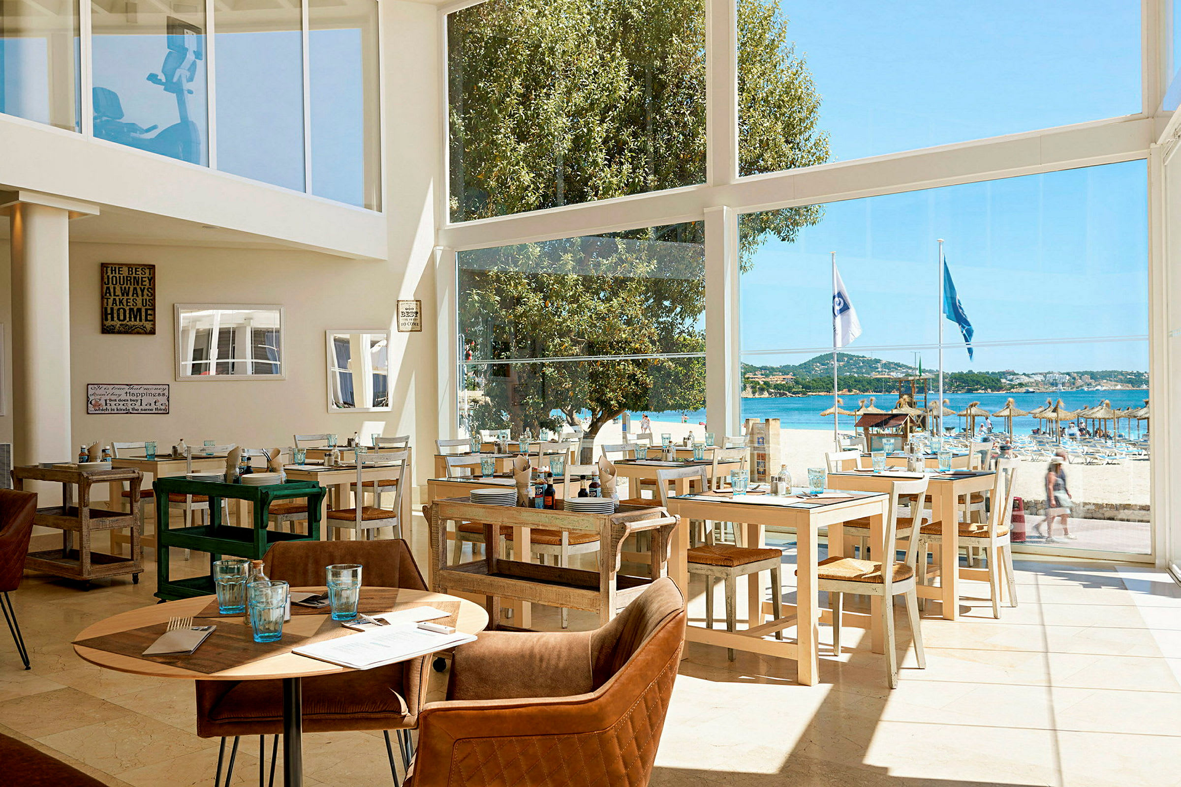 En restaurant med glasvægge, der giver udsigt til en strand med parasoller og blåt hav. (AI-genereret tekst)