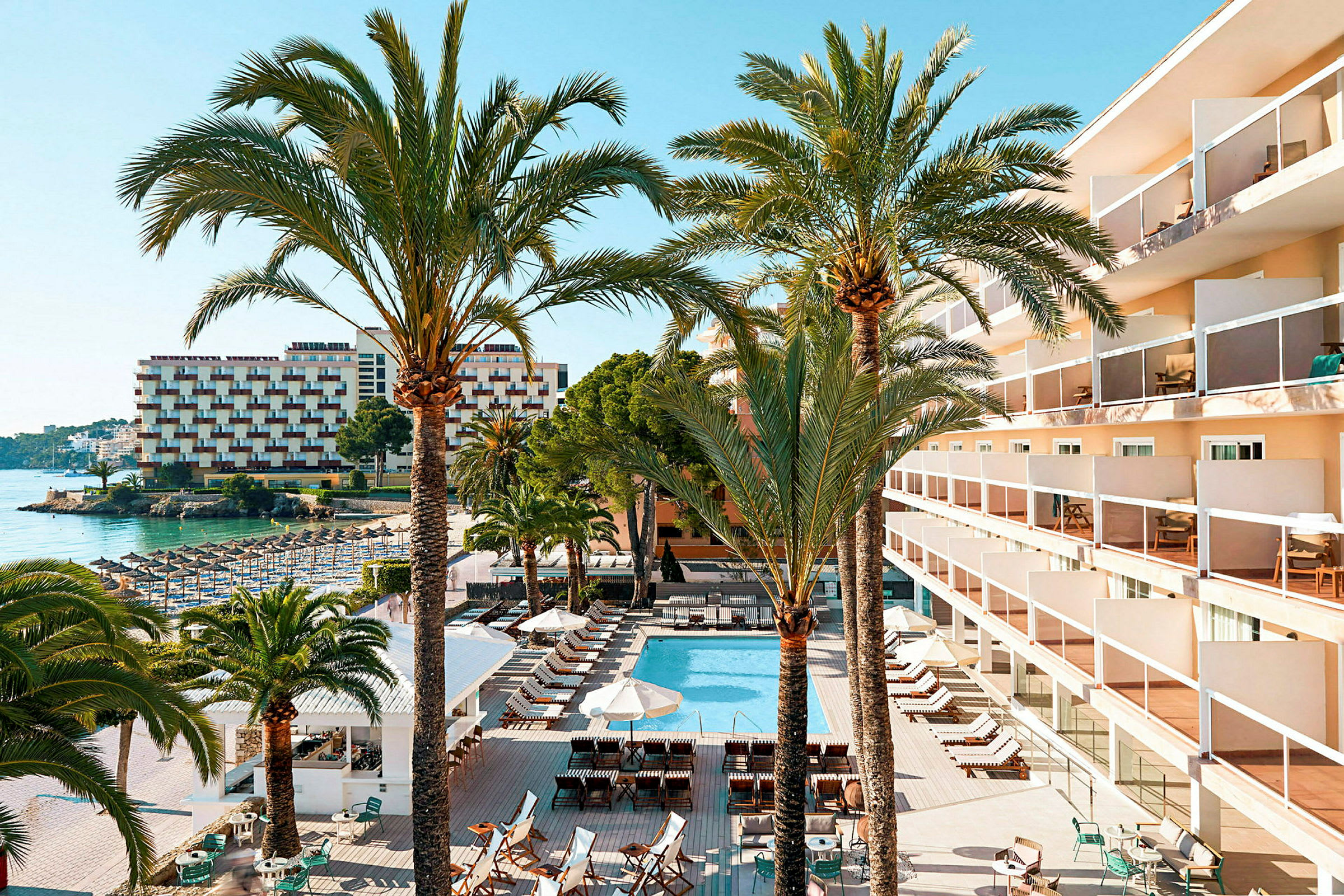 Billede af et hotelområde med en pool, liggestole, parasoller og omkringliggende bygninger. Flere palmer ses i forgrunden. (AI-genereret tekst)