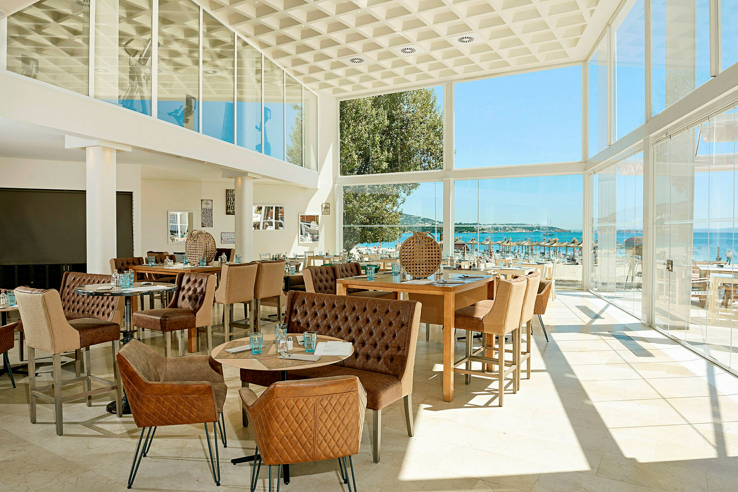Et moderne indrettet restaurantområde med store glasvinduer, der giver udsigt til en strand og havet udenfor. (AI-genereret tekst)