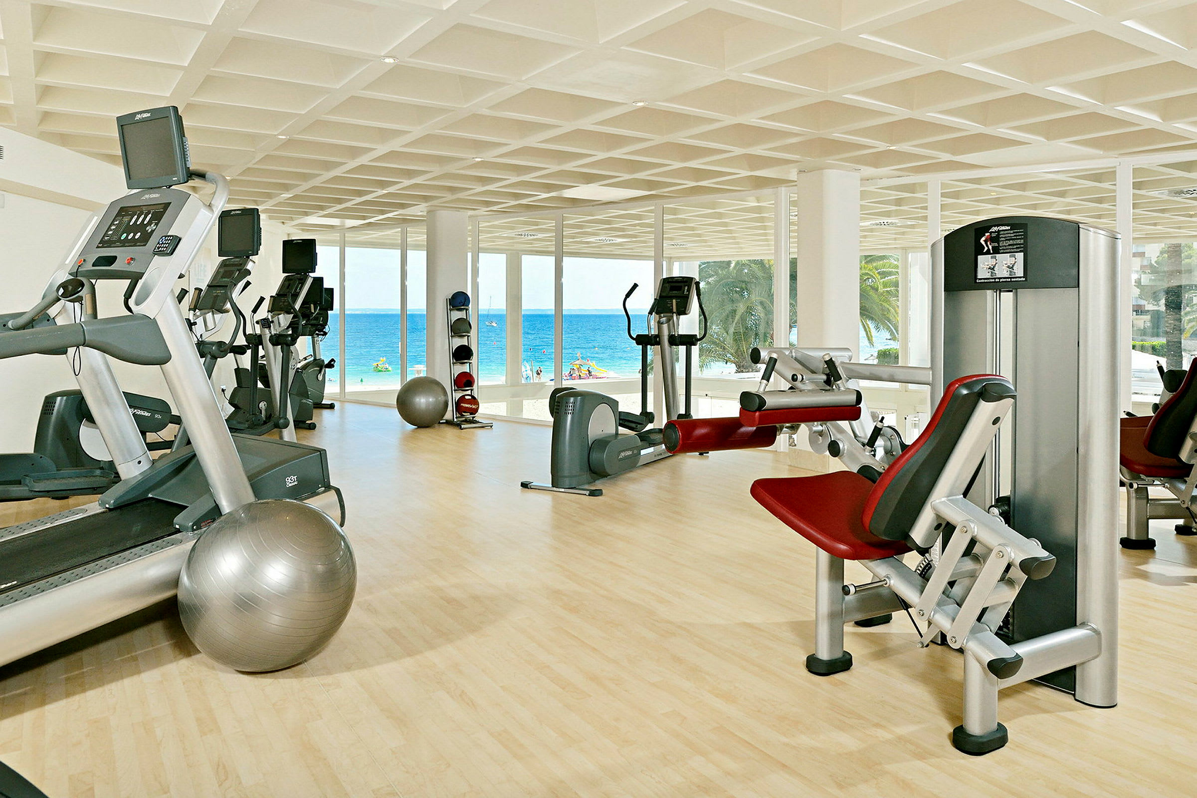 Et fitnesscenter med træningsudstyr og store vinduer ud mod stranden og havet. (AI-genereret tekst)