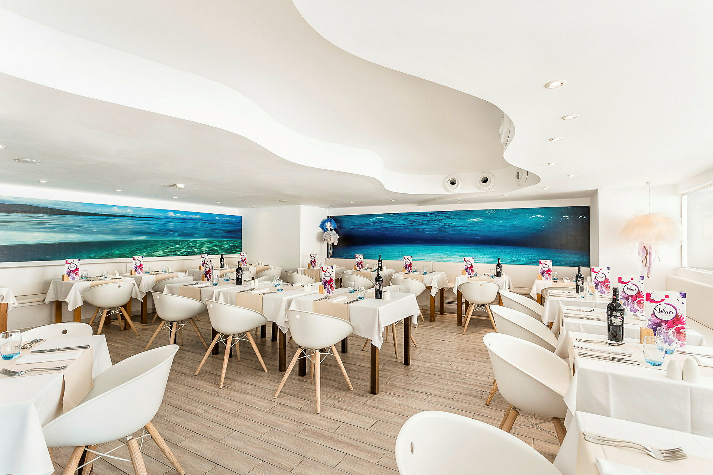 En moderne restaurant med hvide stole og borde samt et stort undervandsmaleri på væggen. (AI-genereret tekst)