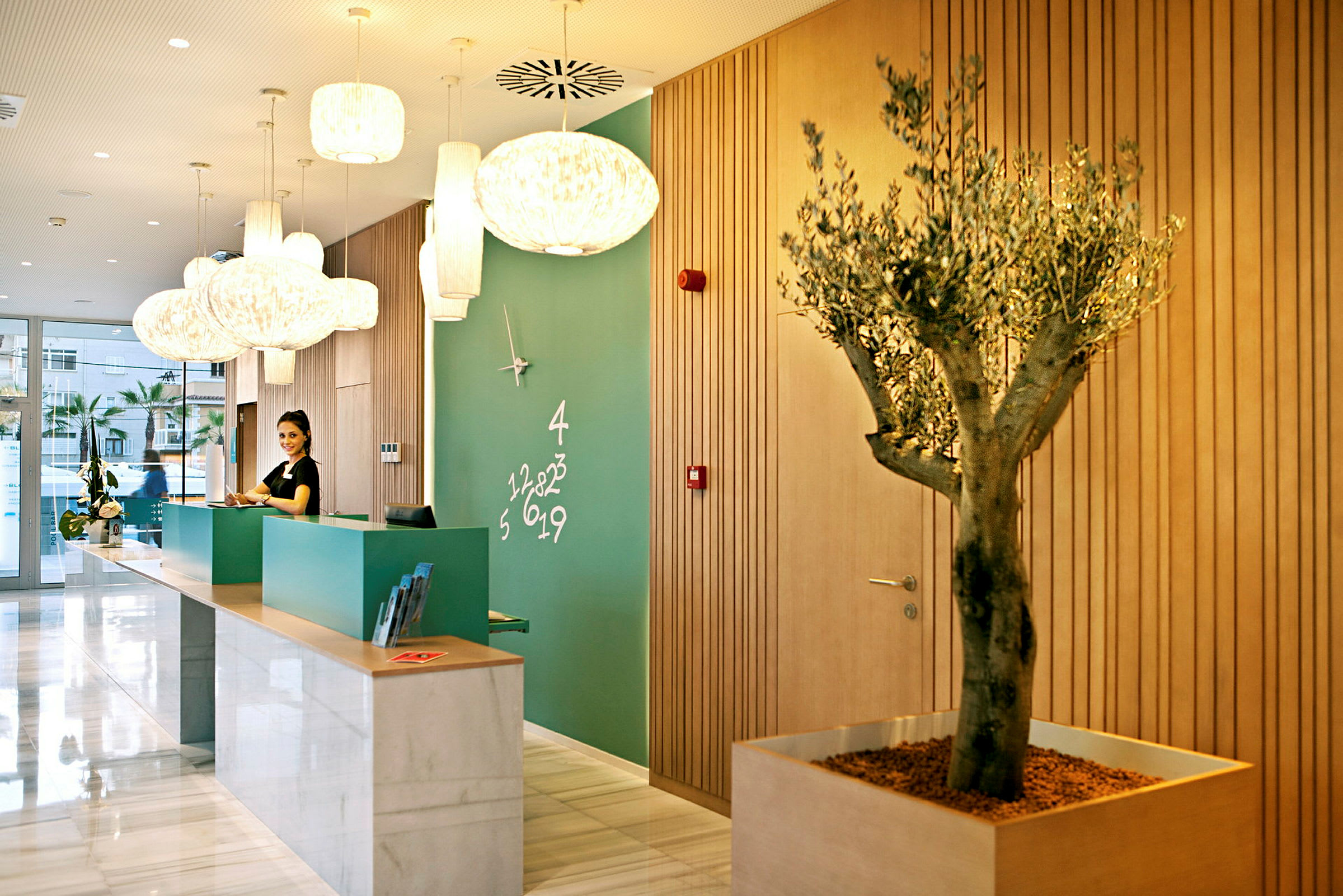 Reception på et hotel med moderne indretning, grøn væg med ur og numerisk dekoration, samt en trækrukke. (AI-genereret tekst)