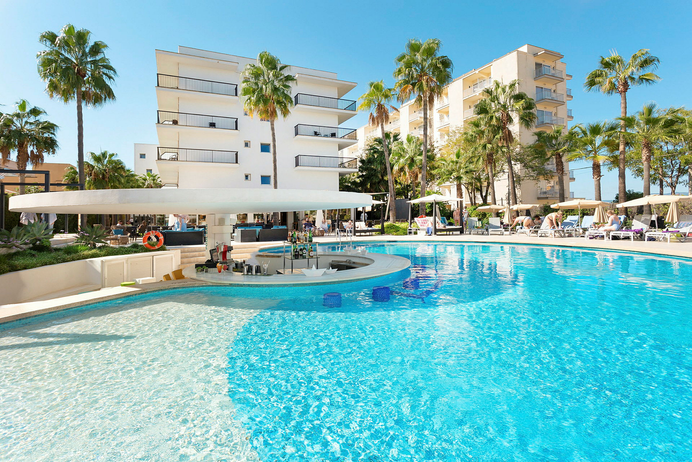 Billede af en pool med bar og omkringliggende liggestole samt bygninger i baggrunden. (AI-genereret tekst)