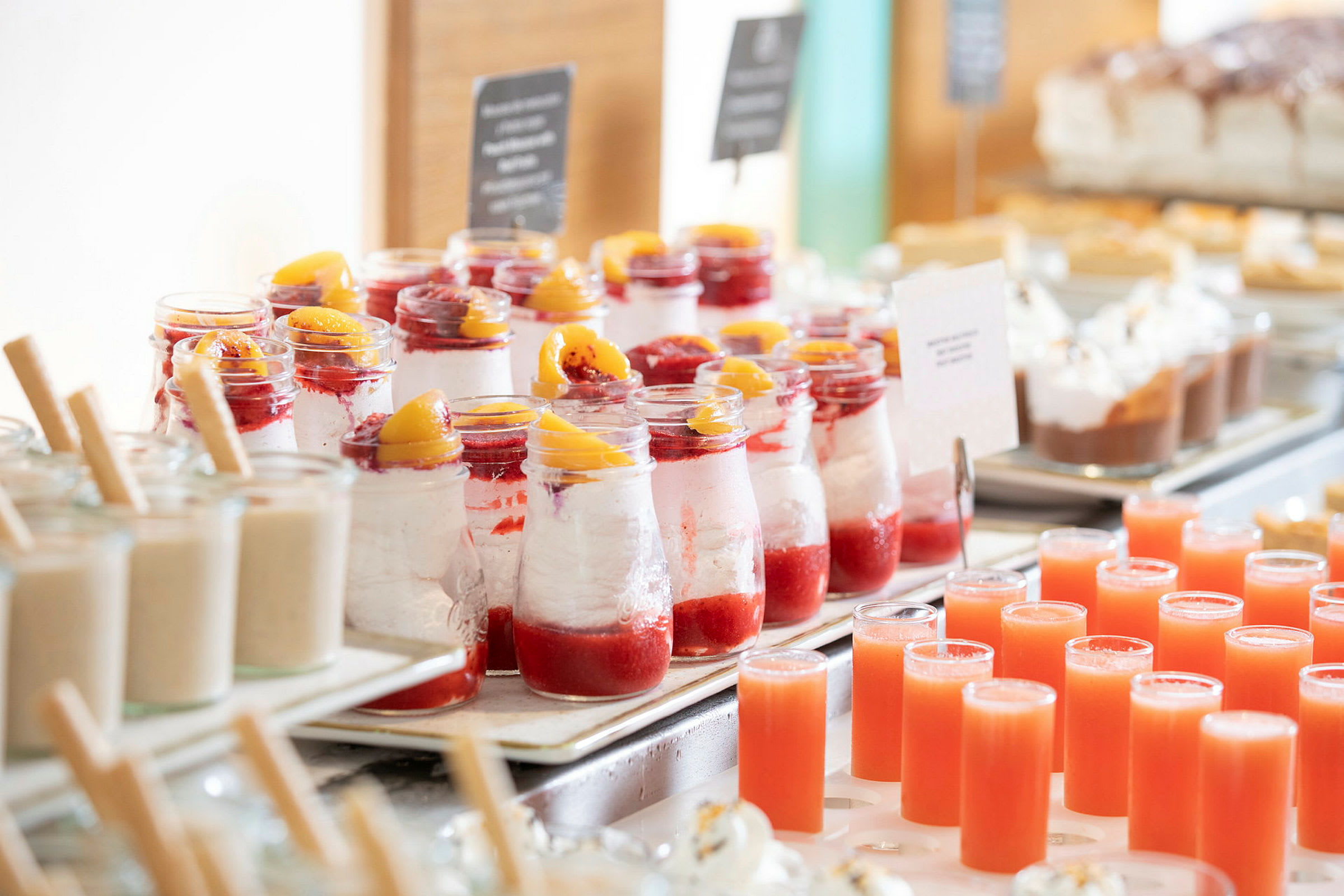 En række af desserter i glas med røde frugtdetaljer og små juiceglas på et bord. (AI-genereret tekst)