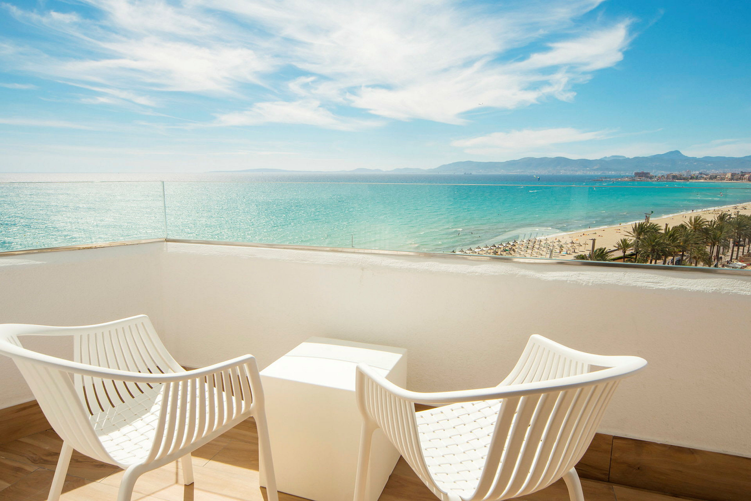 To hvide stole og et bord på en balkon med udsigt over havet og stranden. (AI-genereret tekst)