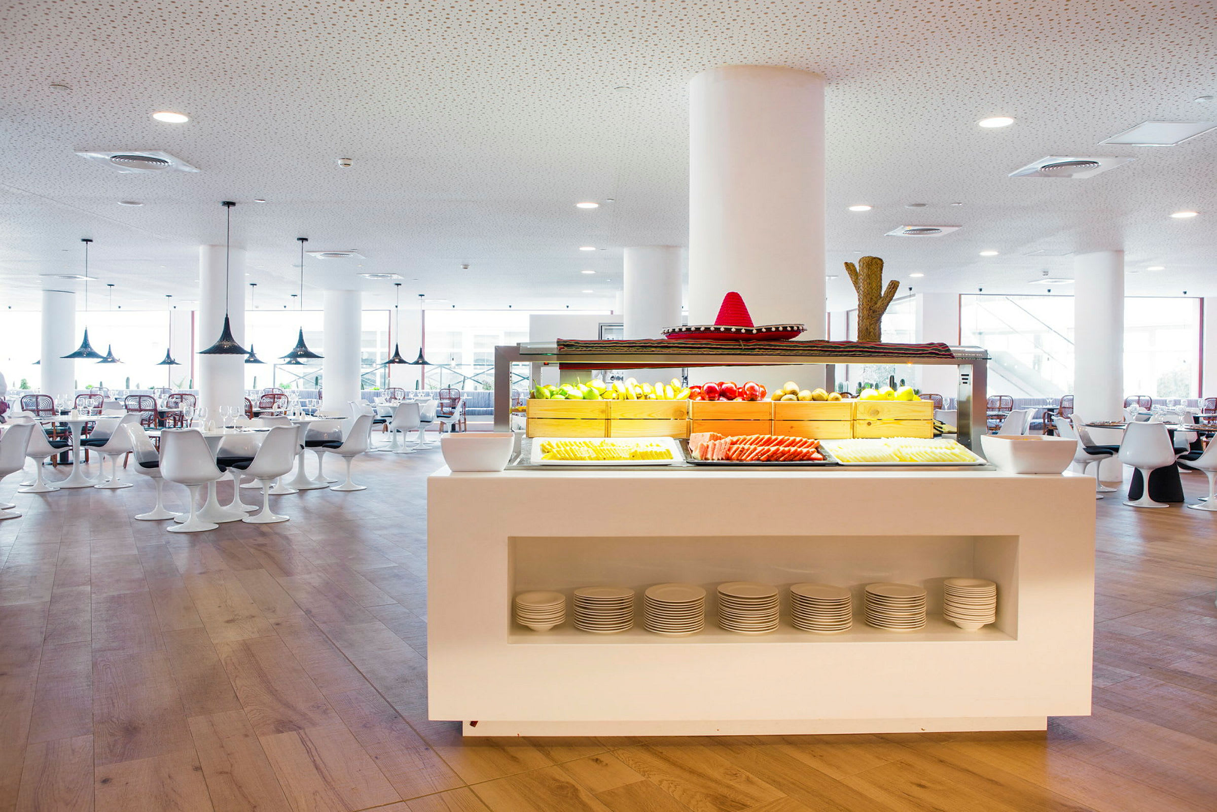 Et restaurantområde med en hvid buffetstation fyldt med frisk frugt og grøntsager, omgivet af moderne hvide borde og siddepladser. (AI-genereret tekst)