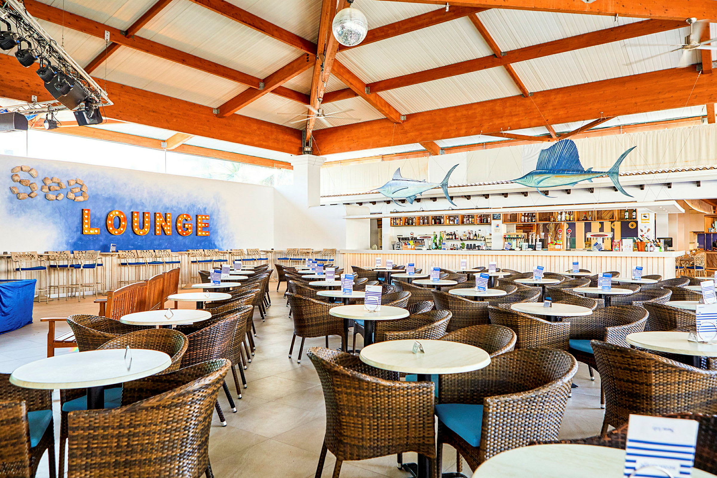 Interiør af en strandbar med rattanstole og borde, belyst tekst med ordene 'Sea lounge' på væggen. (AI-genereret tekst)