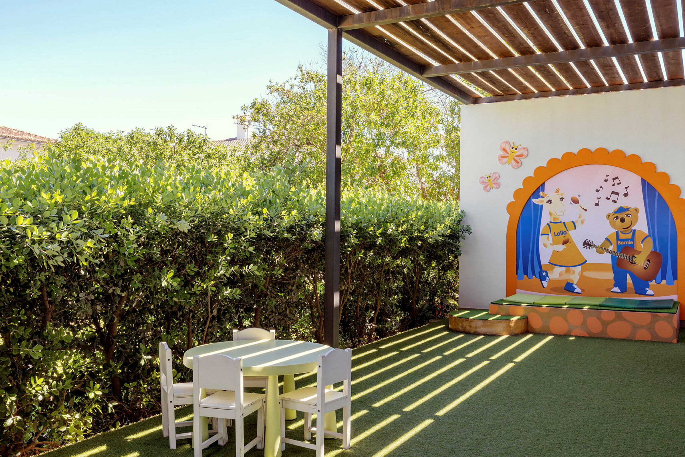 En udendørs legeplads med et farverigt vægmaleri, der viser to figurer, samt et bord og stole lavet af træ under en træpergola. (AI-genereret tekst)