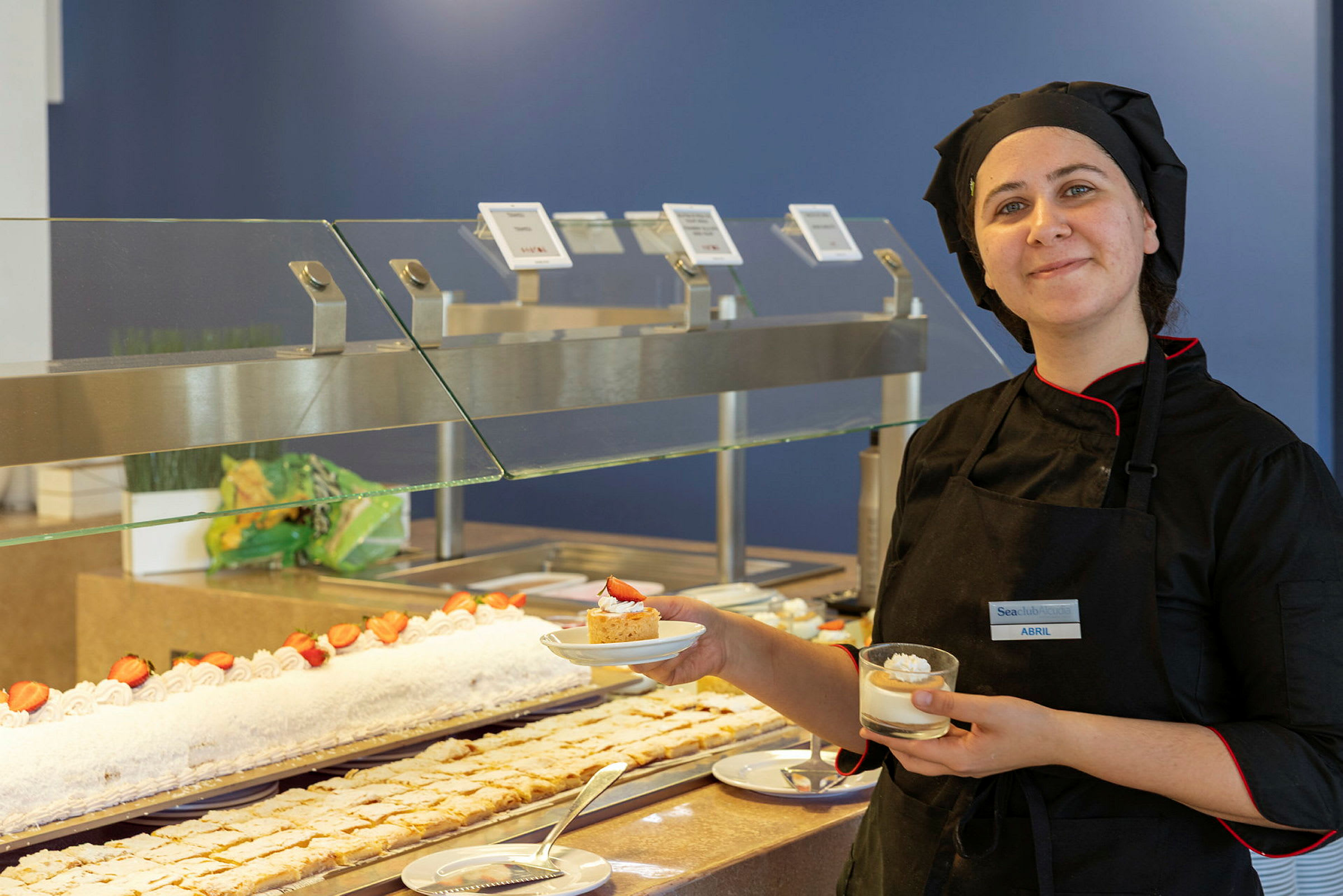 En kok klædt i sort står ved en buffet med desserter og holder to serveringer. (AI-genereret tekst)