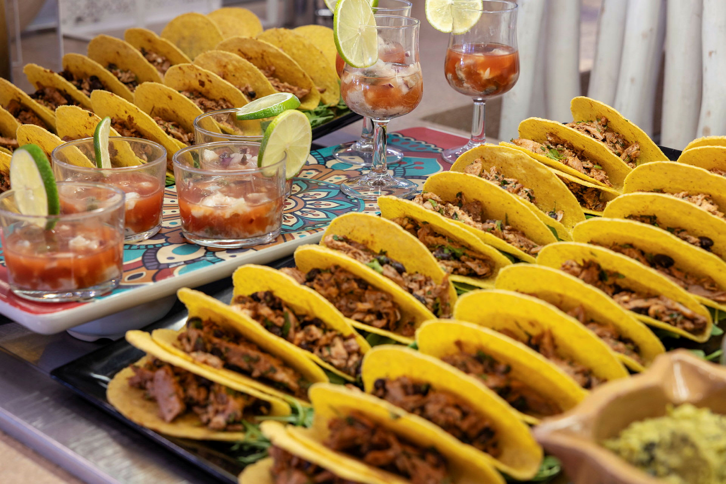Billede af flere tacos fyldt med kød og grøntsager, serveret på bakker sammen med glas med drikke og limebåde. (AI-genereret tekst)