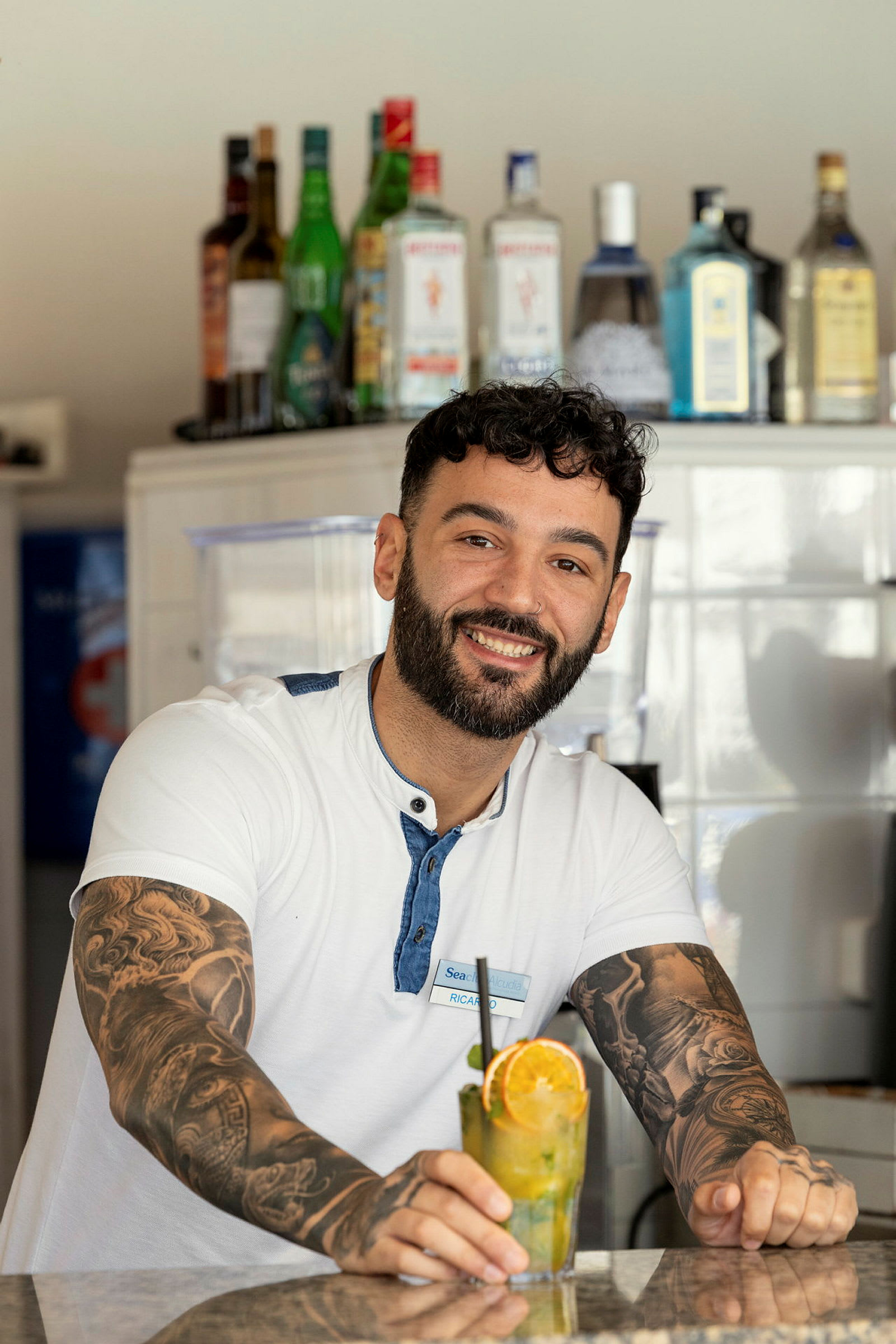 En smilende mand står ved en bardisk med forskellige spiritusflasker i baggrunden og holder en cocktail med appelsinskive. (AI-genereret tekst)