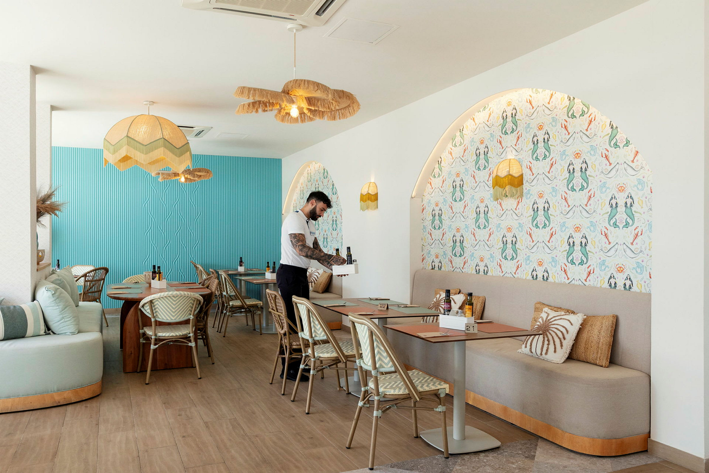 En interiør af en moderne restaurant med blå og mønstrede vægge, borde med stole og en tjener, der forbereder en plads. (AI-genereret tekst)